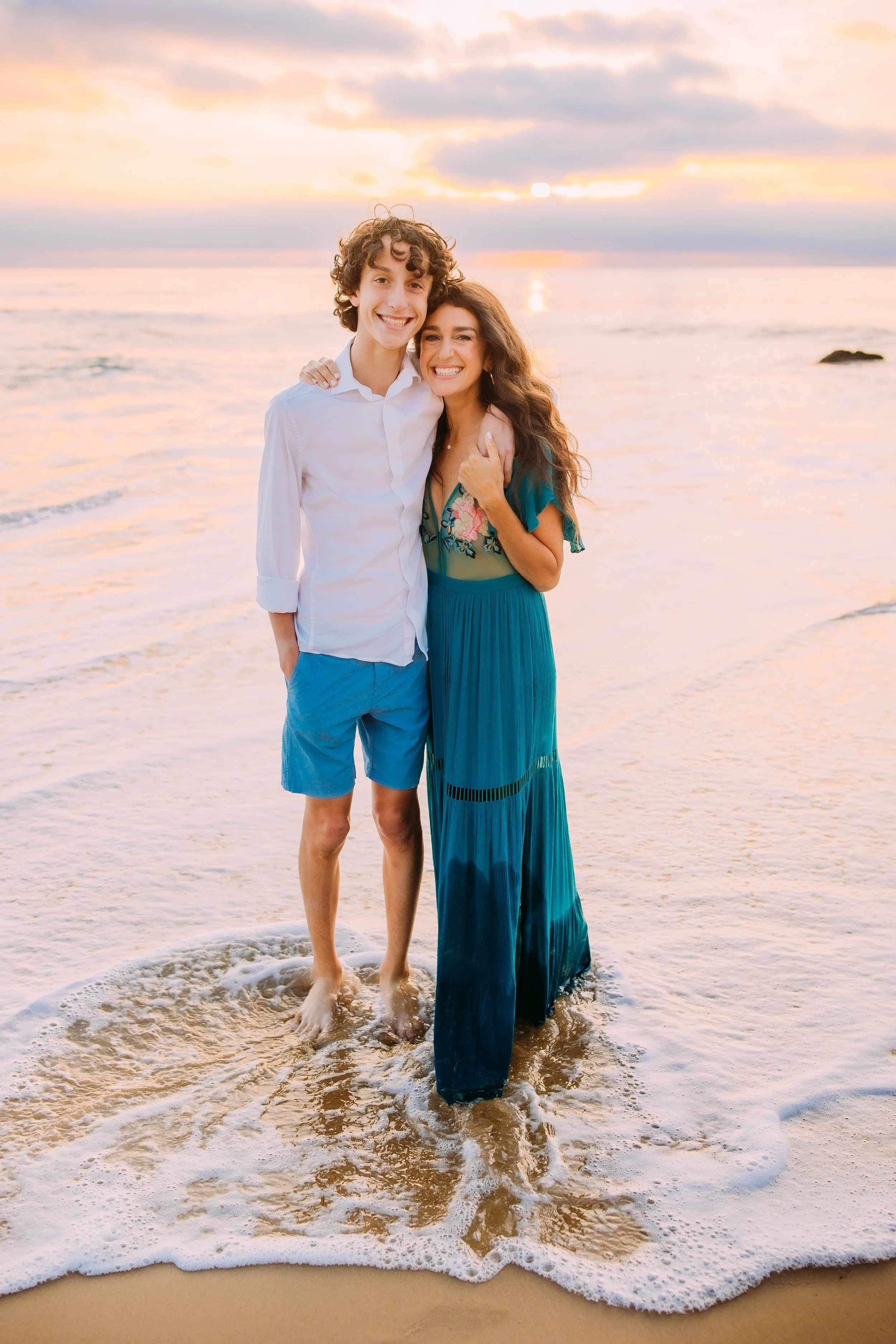 mom and son smiling at the camera while standing in the ocean tide during their photo shoot in Newport Beach, CA