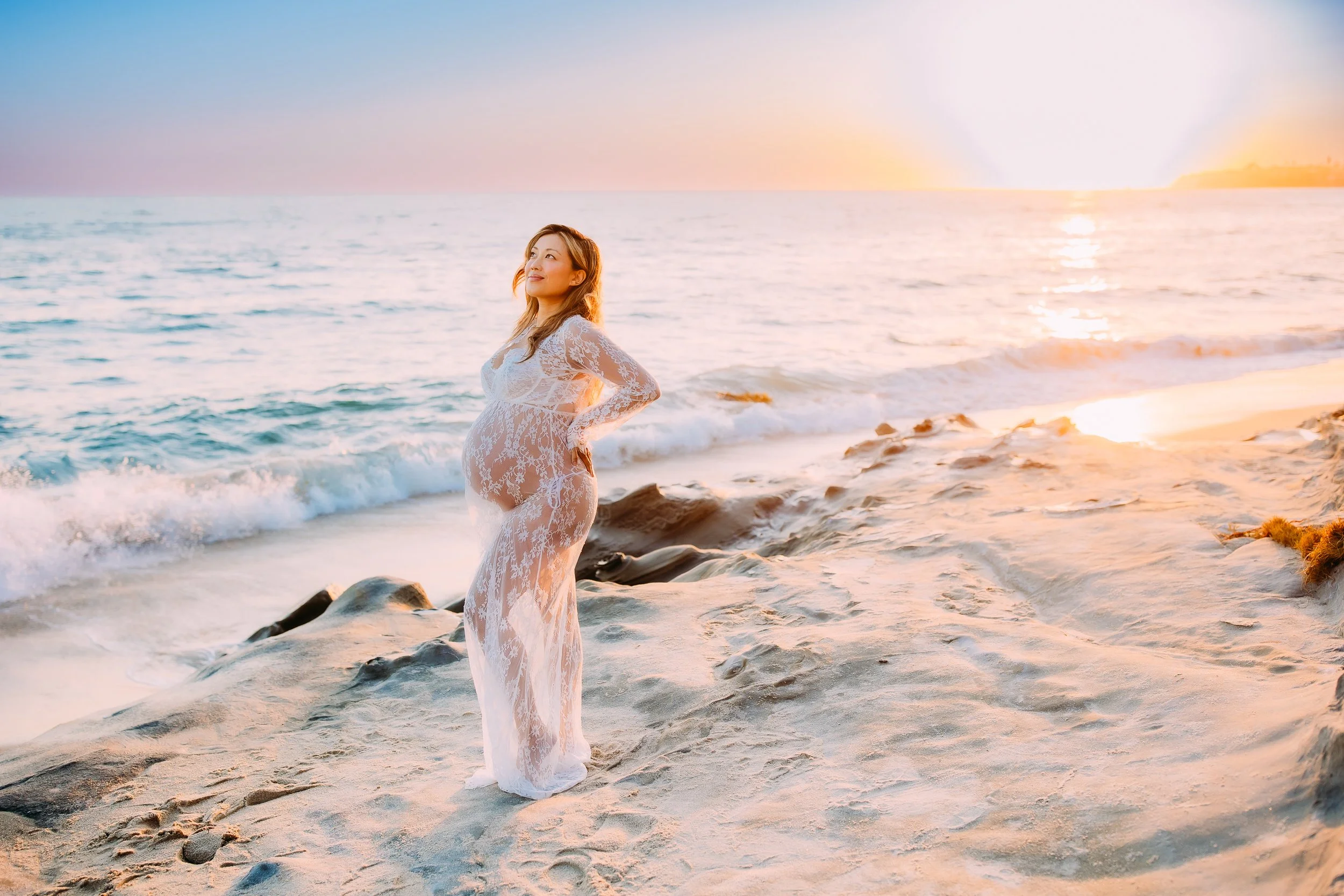 pregnant mother to be gazing upward with her hand resting on her back and the sun setting behind her during her maternity photoshoot in Laguna Beach, California