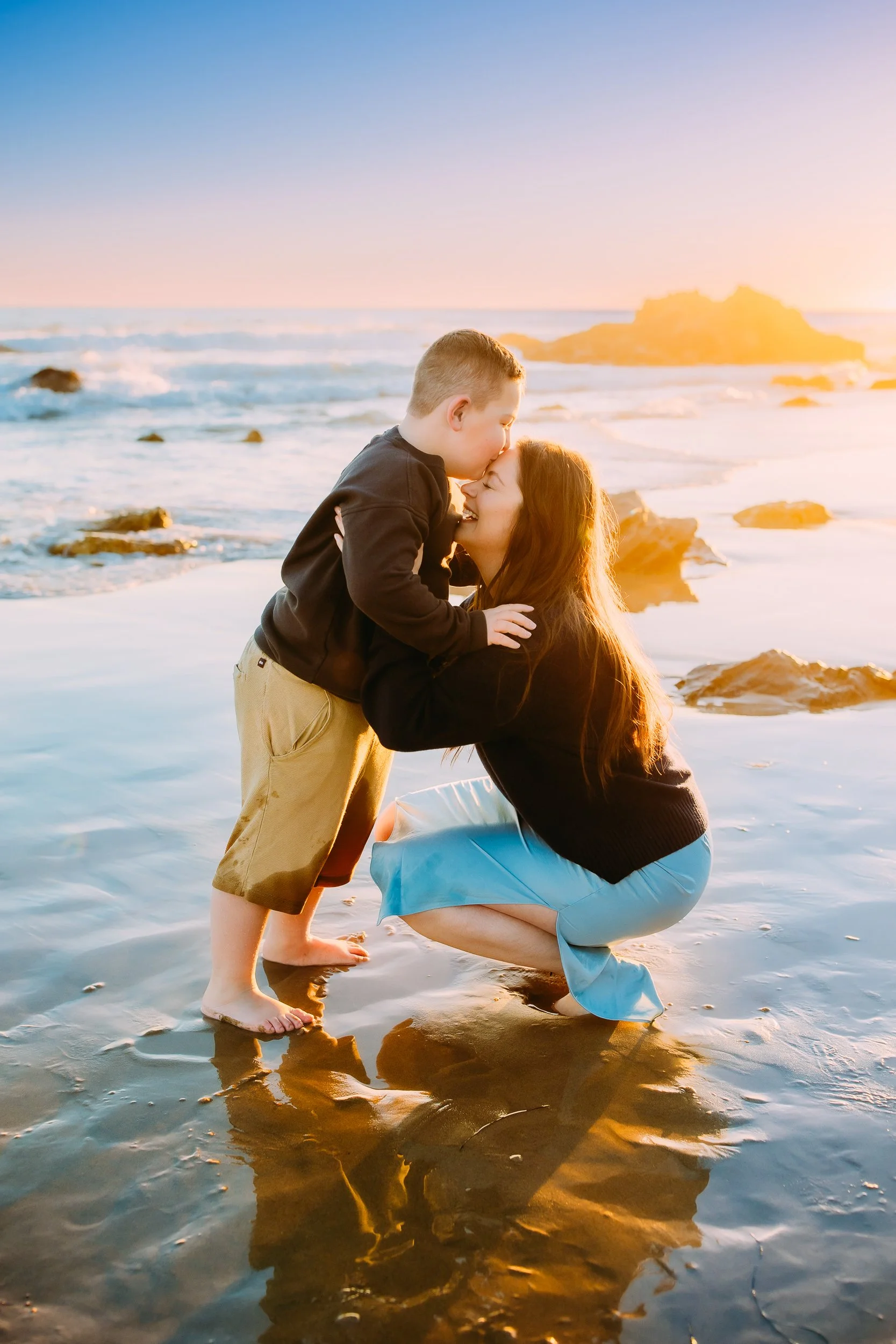 son playfully kissing his laughing mom on her forehead while the sun sets behind them during their ocean photoshoot in Newport Beach.