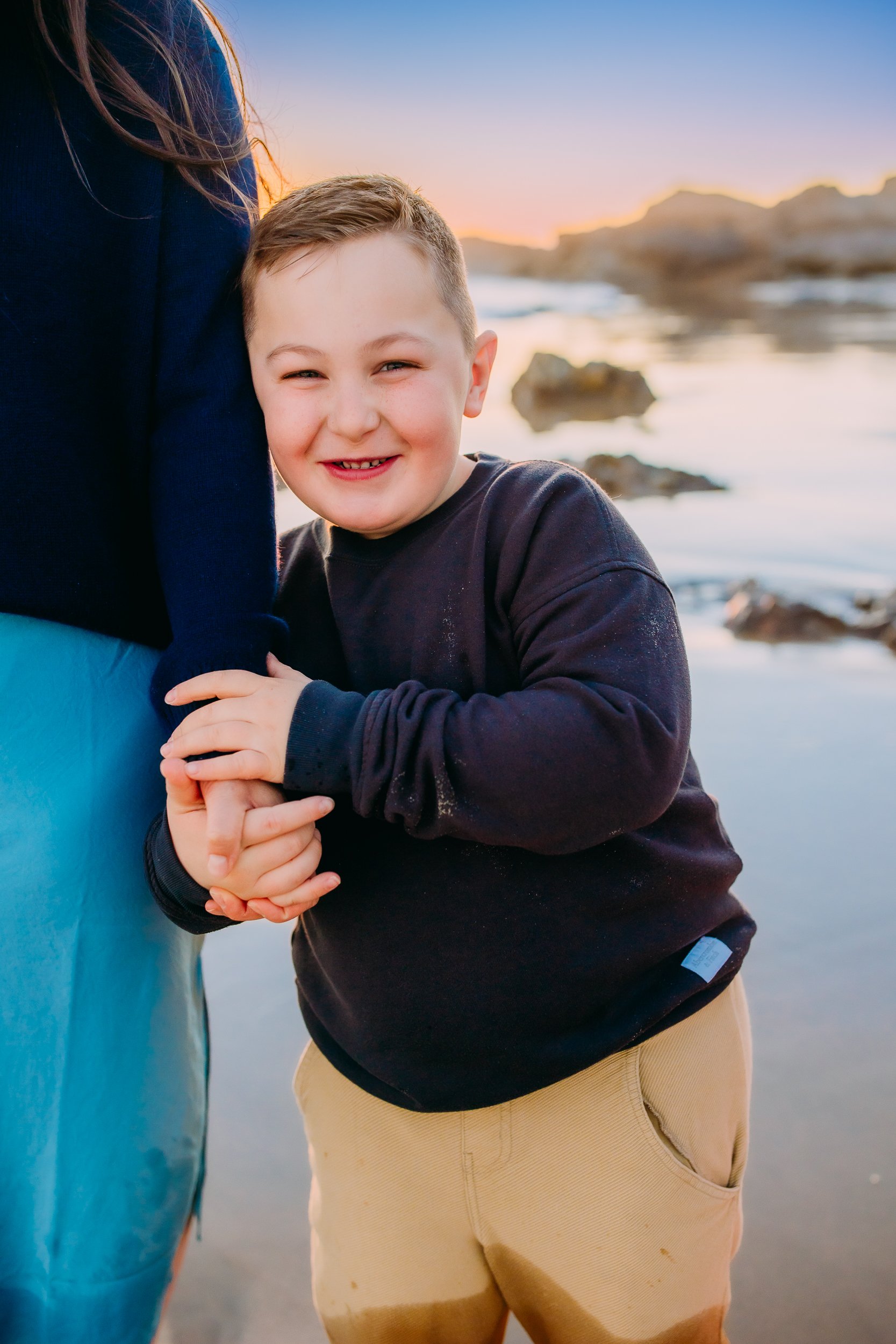 close up image of boy smiling at the camera while holding his mom's hand during their sunset beach shoot in Newport Beach.