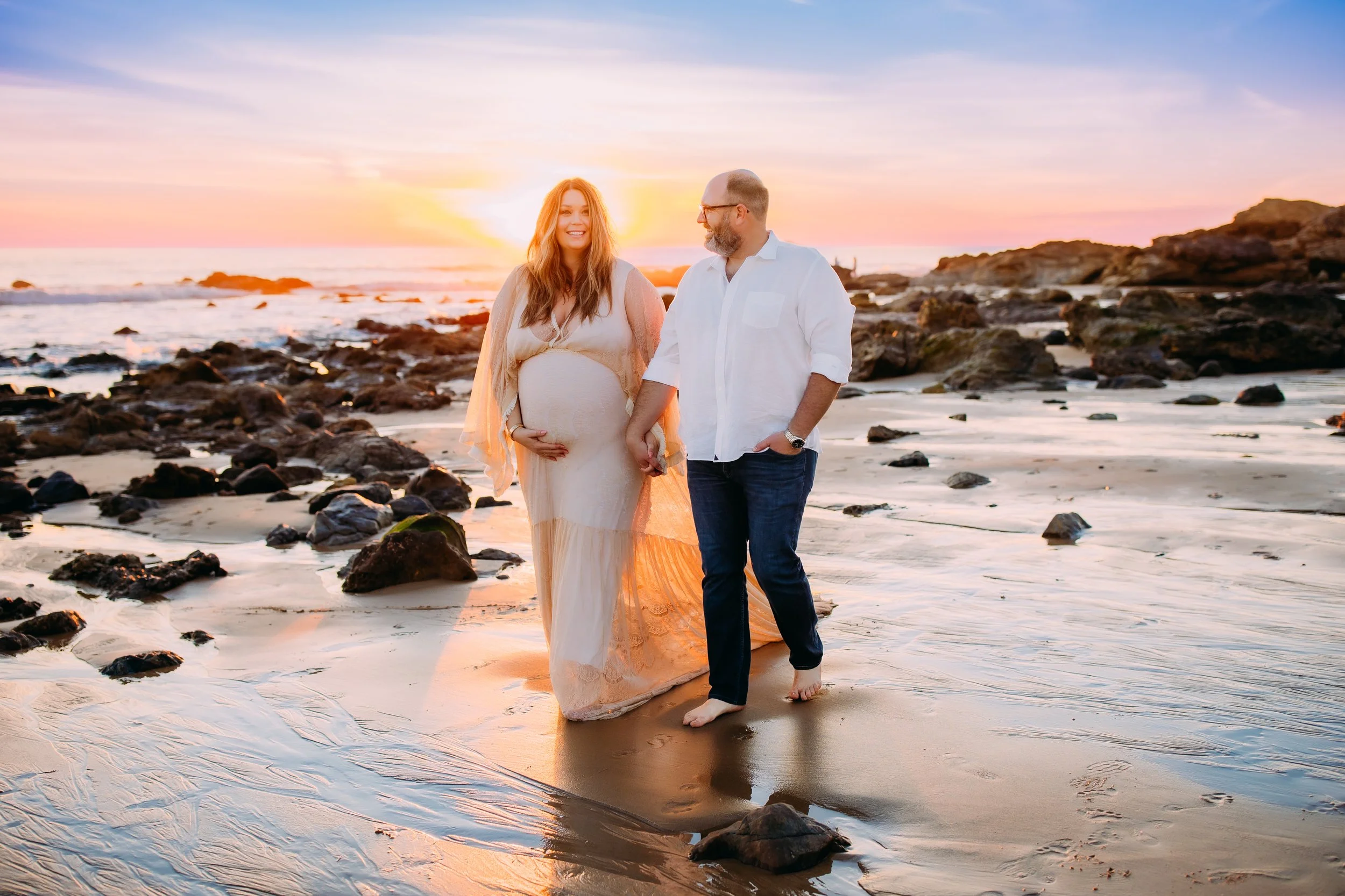 husband and wife holding hands as they walk along the shoreline at sunset during their photo session in Newport Beach Orange County California