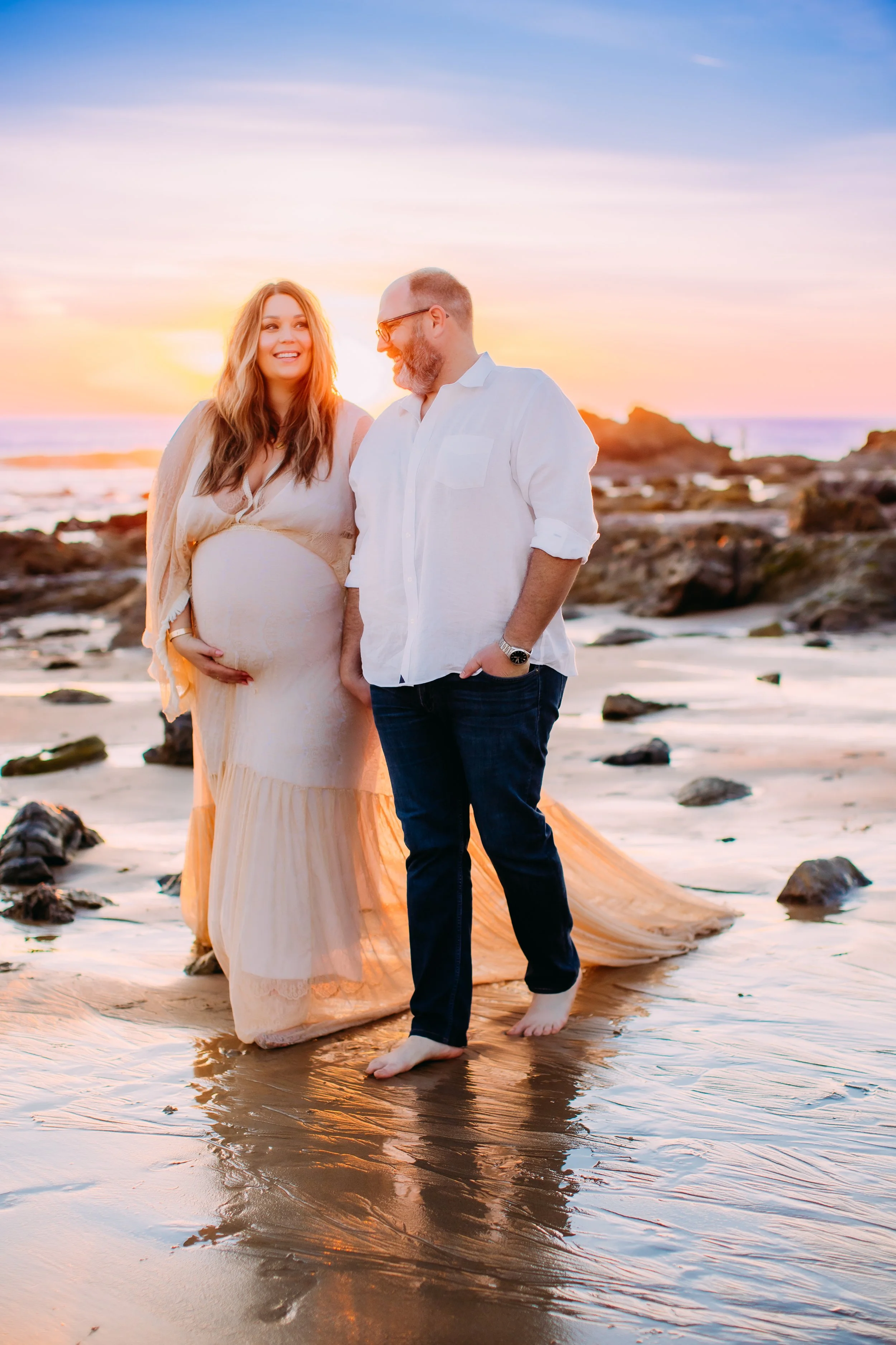 dad and mom to be holding hands and smiling with each other while walking along the shoreline during their photoshoot in Newport Beach, California