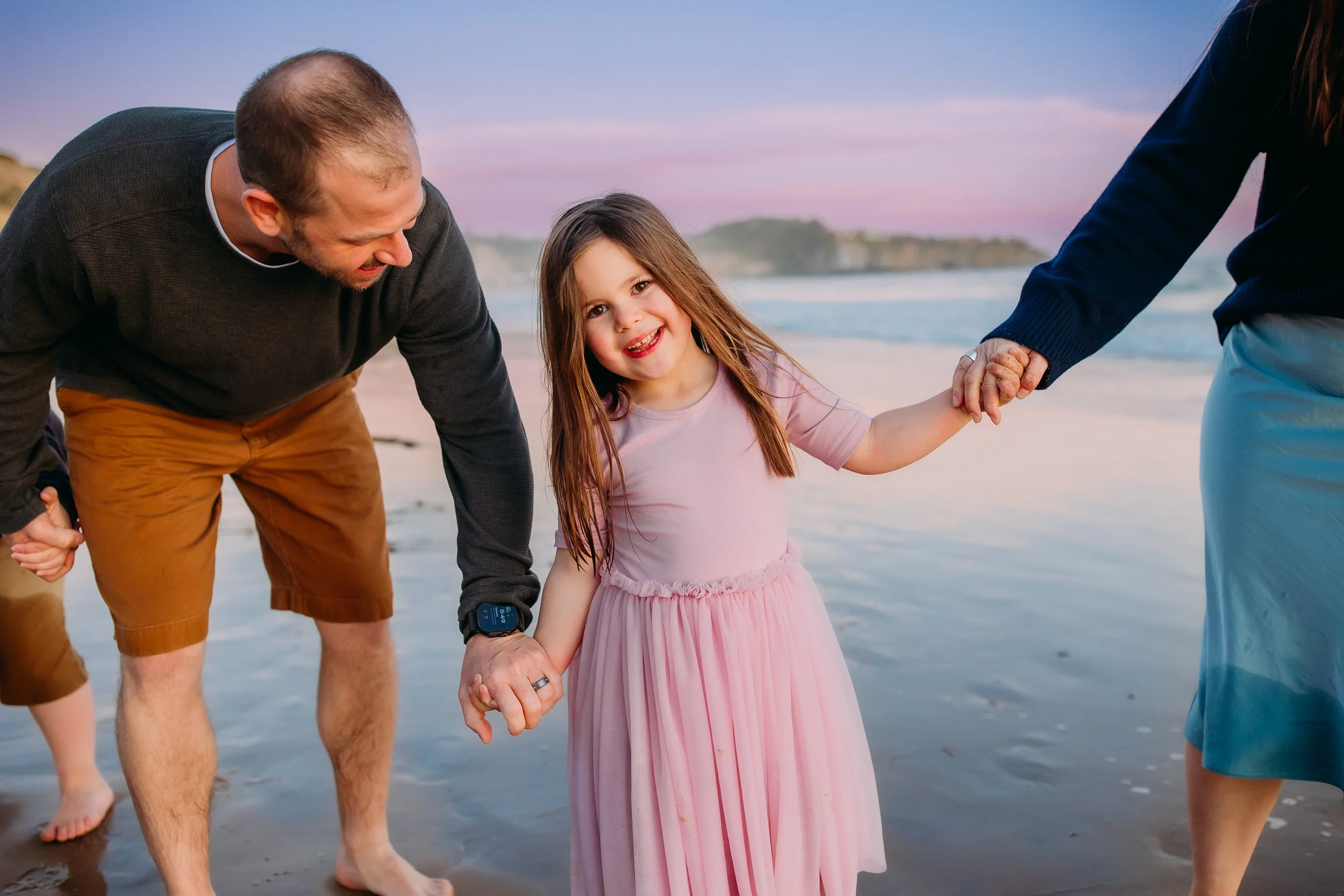 family holding hands and laughing while daughter smiles at the camera during their beach photo session in Newport Beach.