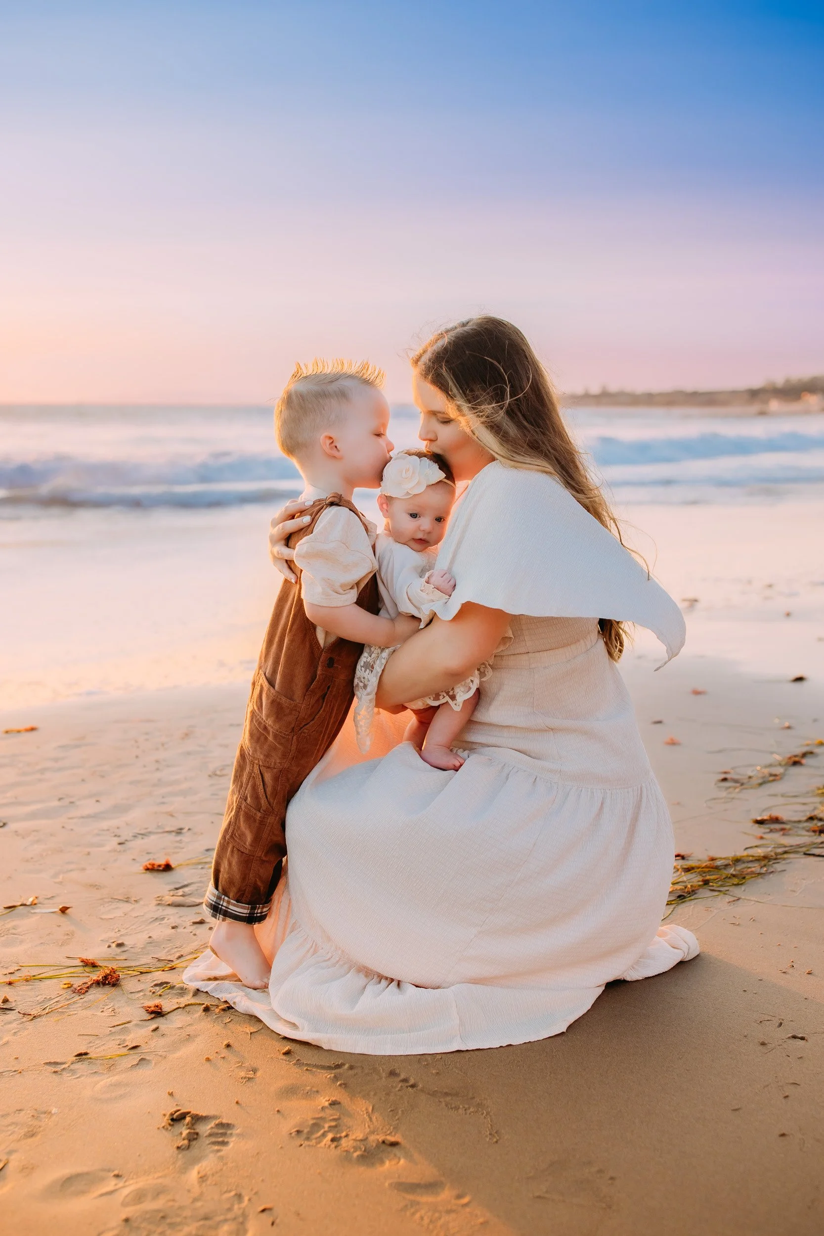 family photo session in Newport Beach, CA where mom and son are kissing their baby sister at sunset on the beach