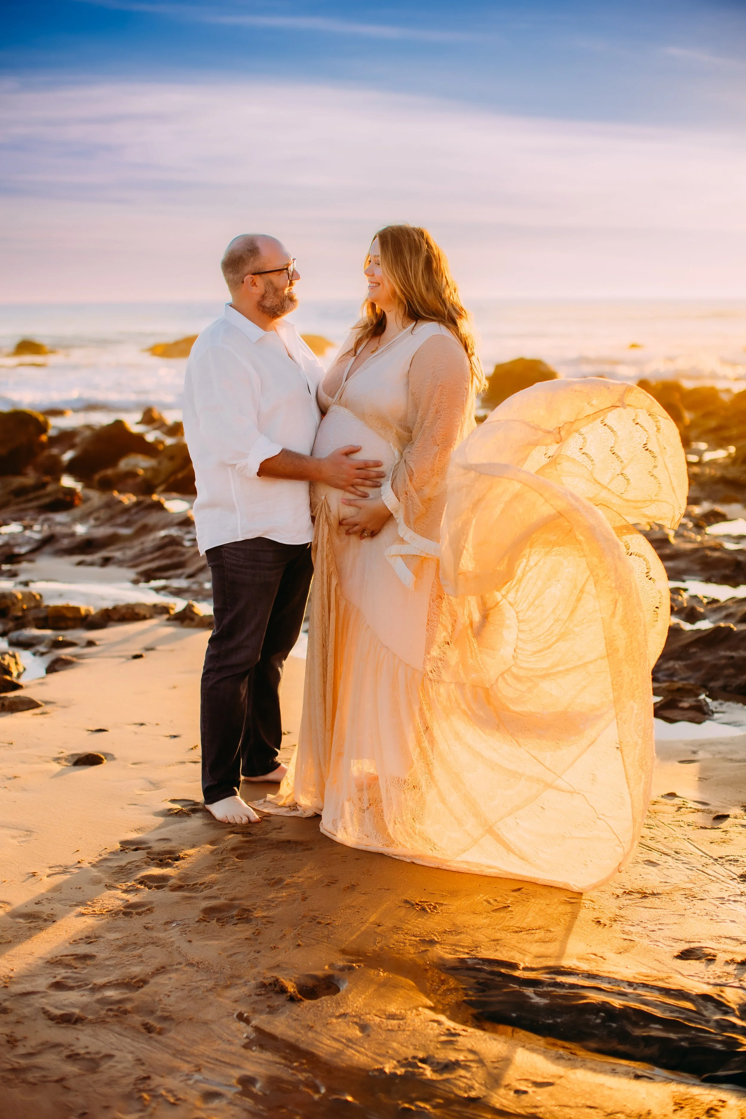 pregnant woman and her husband smiling at each other with hands on her belly while her dress blows into the air during the photo session in Newport Beach, California