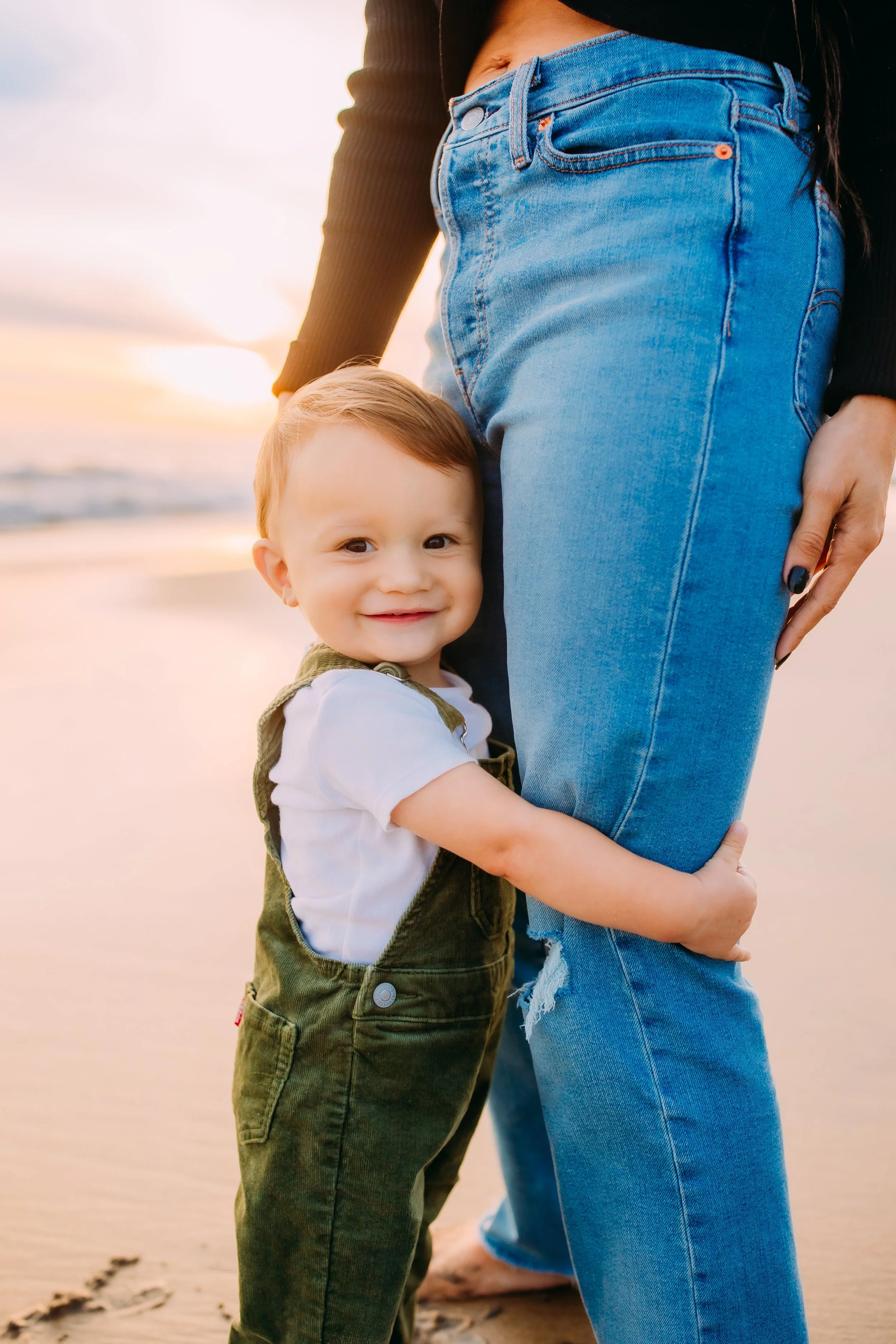 one year old holding onto his mom's leg on the beach during a Newport Beach mommy and me photoshoot.