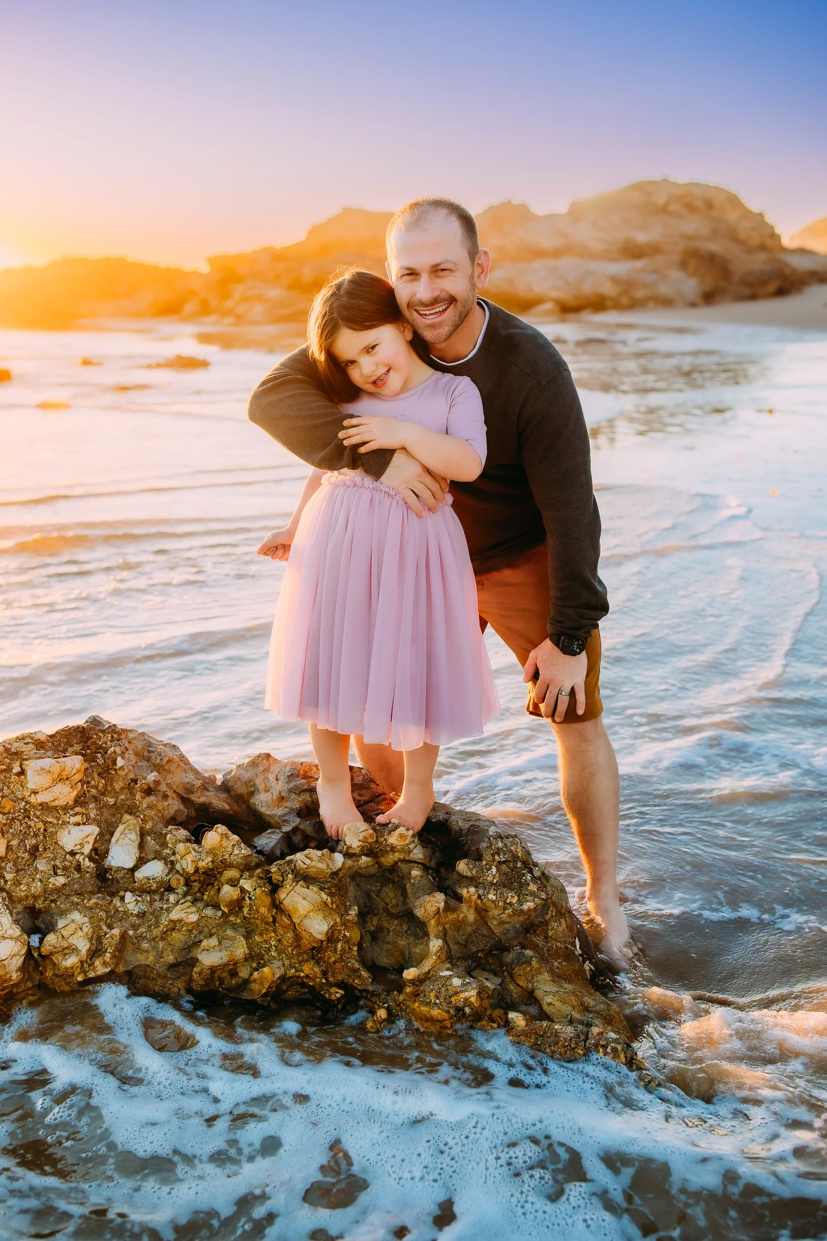 dad and daughter joyfully posing for the camera with the sun setting behind them during their family photos in Newport Beach.