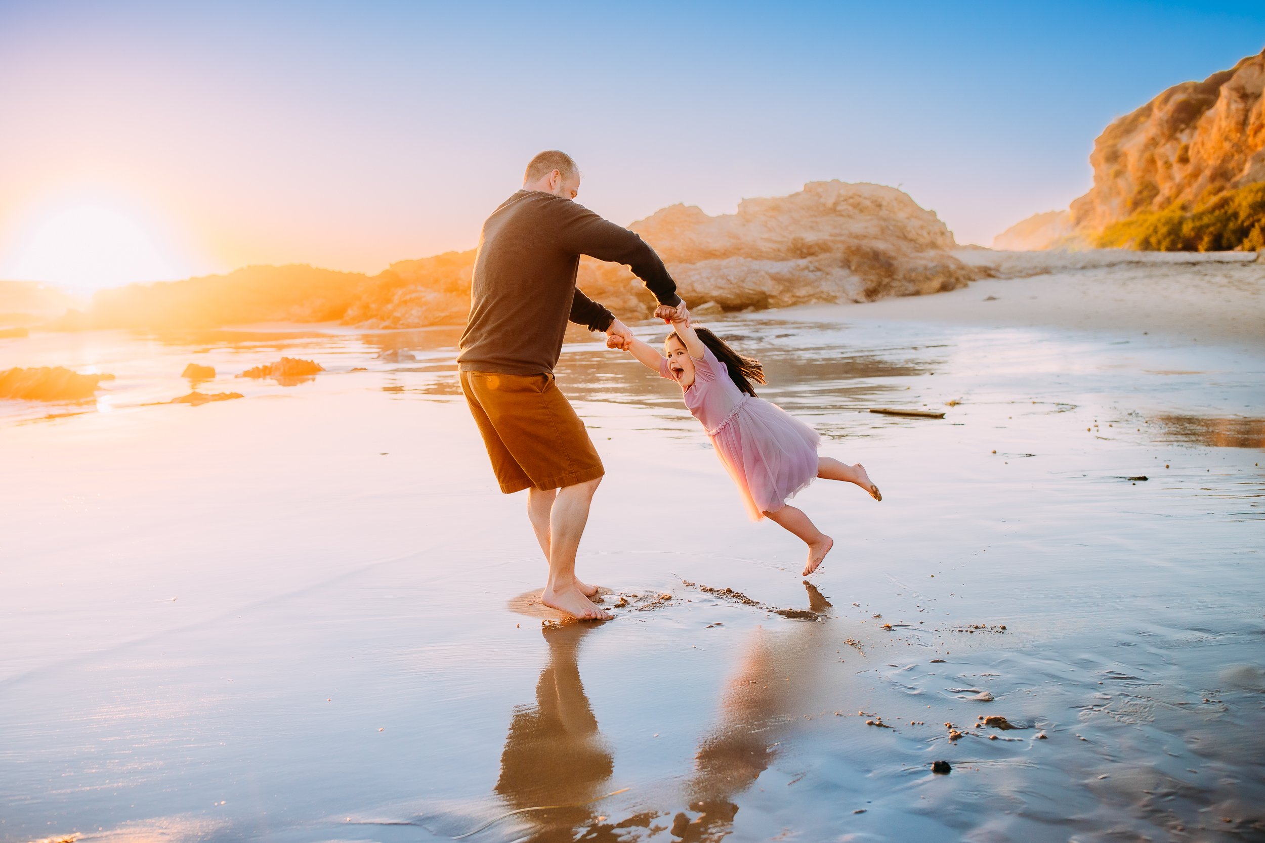 dad swinging his daughter by her arms during their family photoshoot at a sun soaked sunset in Newport Beach.
