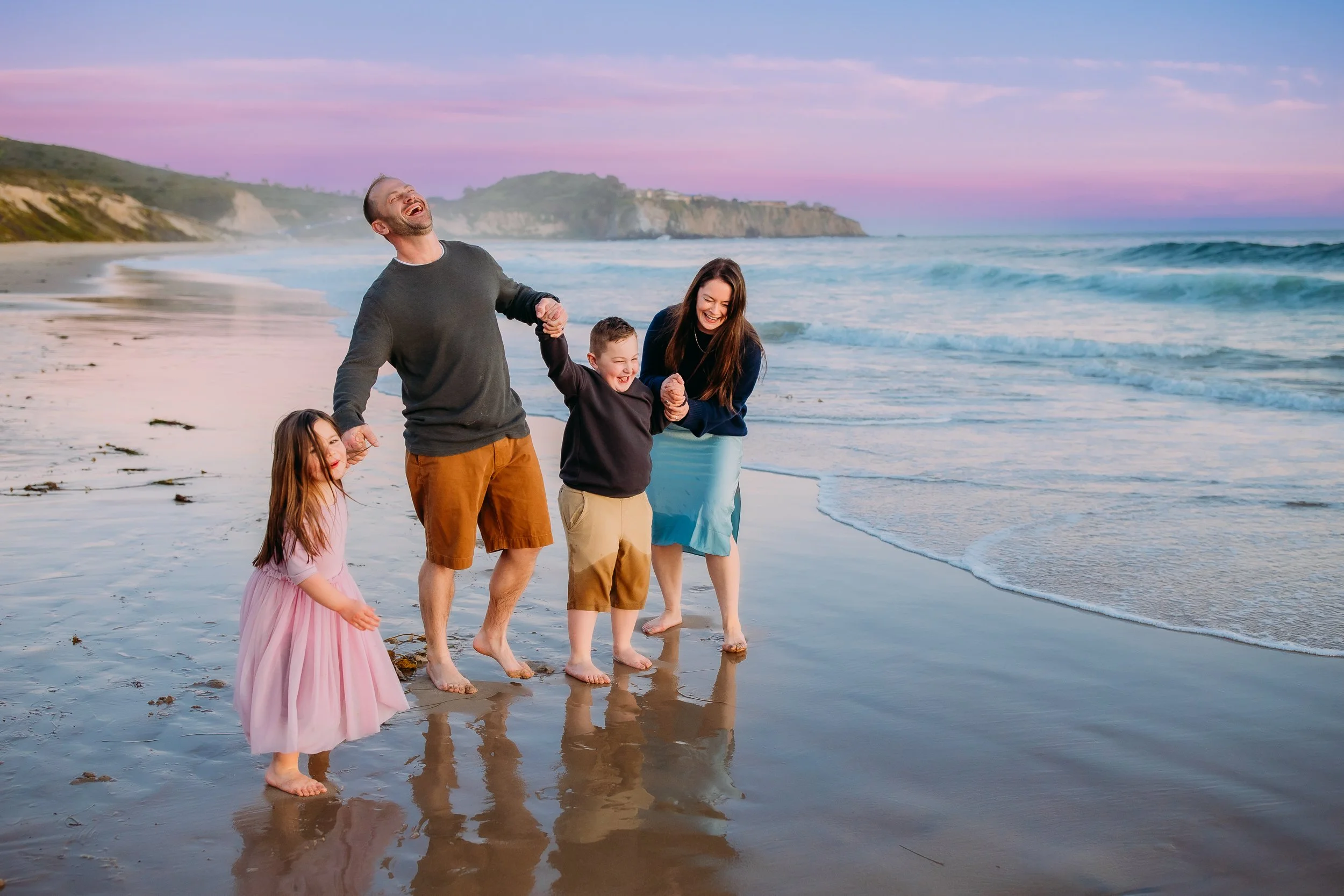 family all laughing together while holding hands at the ocean tide after the sun set during their family photo session in Newport Beach.
