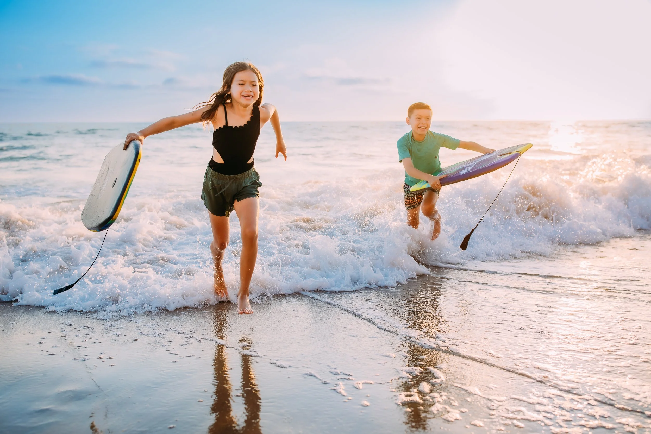 siblings playing in the waves with their boogie boards at sunset during their family photos in Newport Beach, California