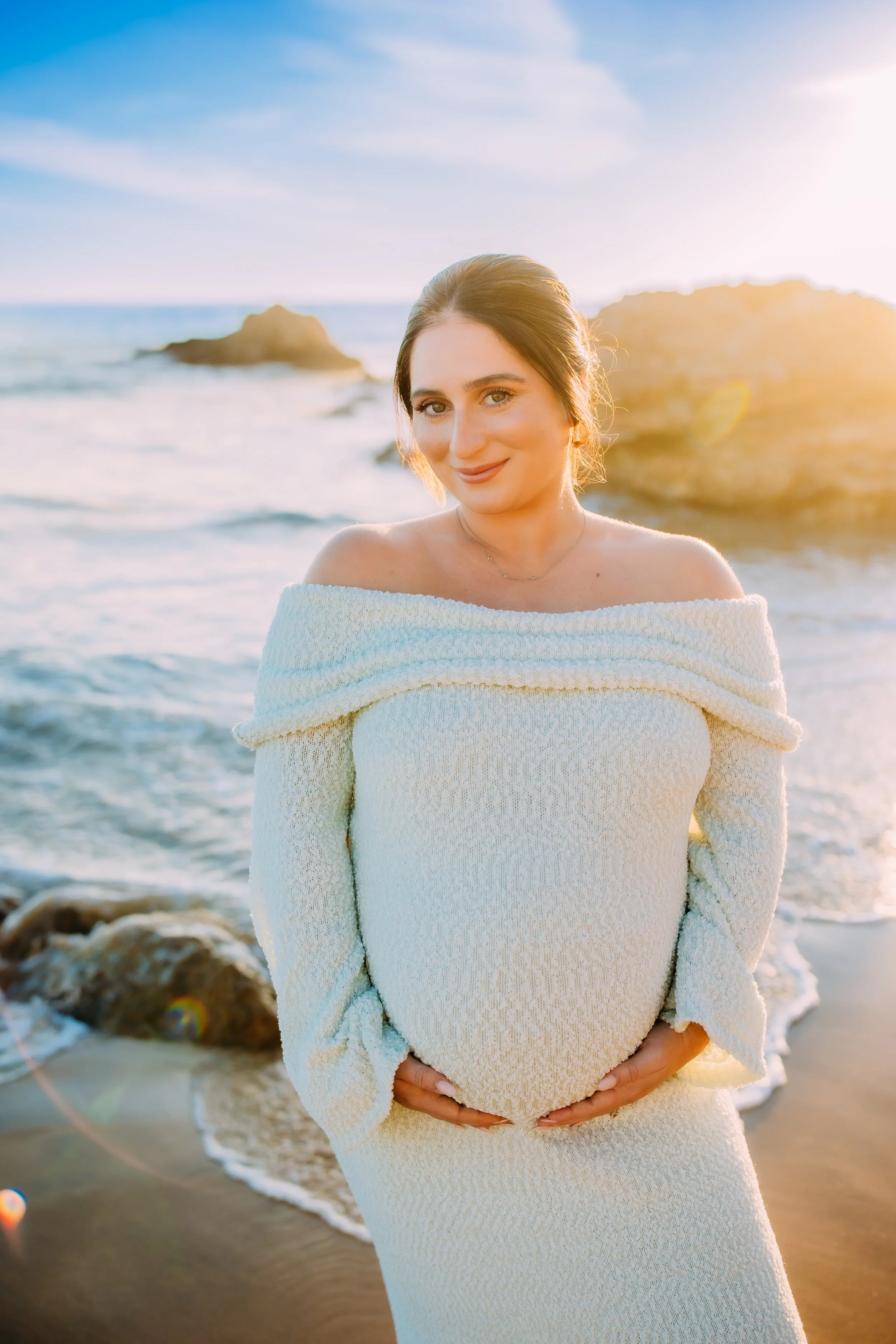 mom to be holding her belly and smiling at the camera during her photoshoot at the ocean in Orange County, Newport Beach, California