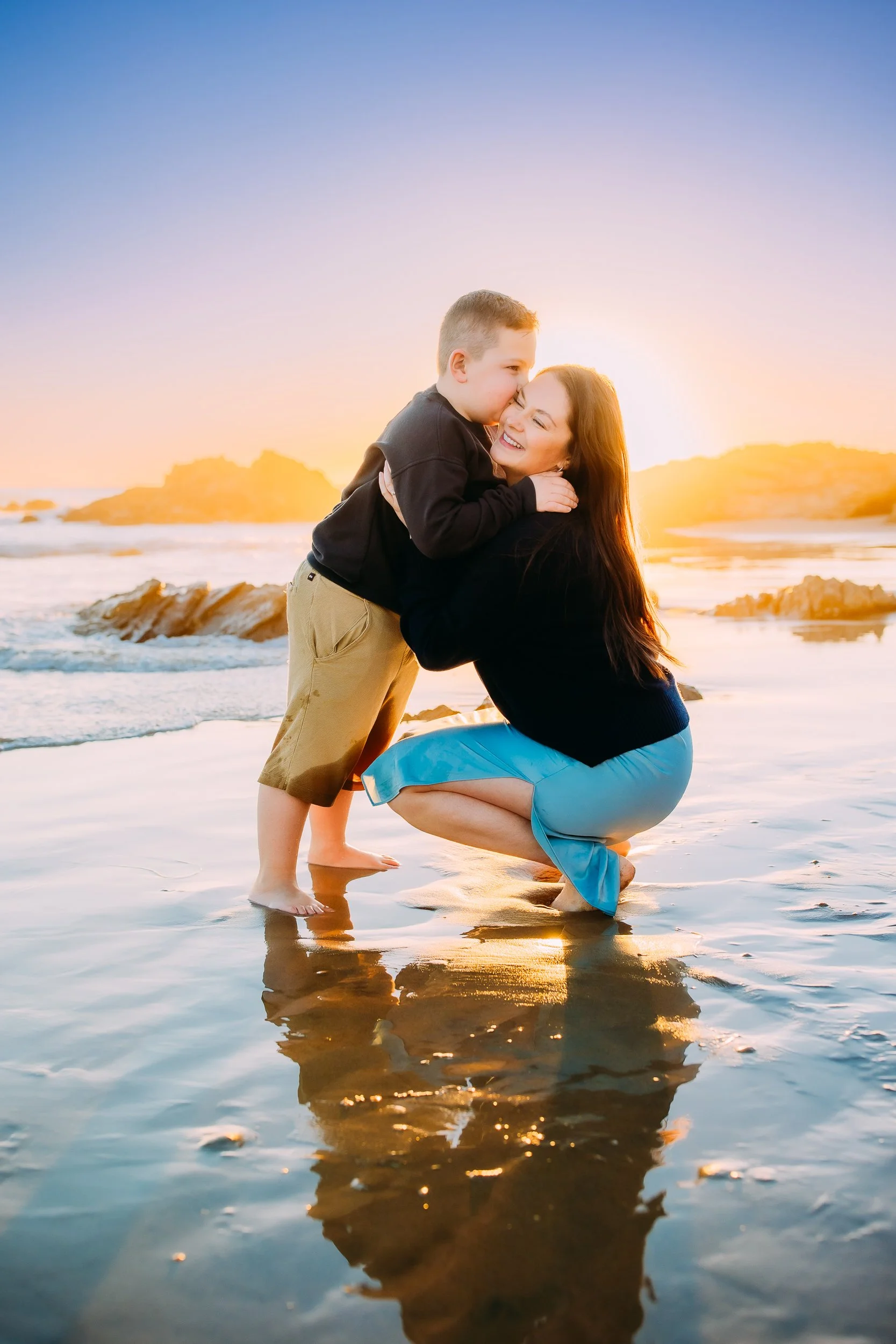 son sweetly kissing his mom while she joyfully smiles with the sun setting behind them during their family photoshoot in Newport Beach.