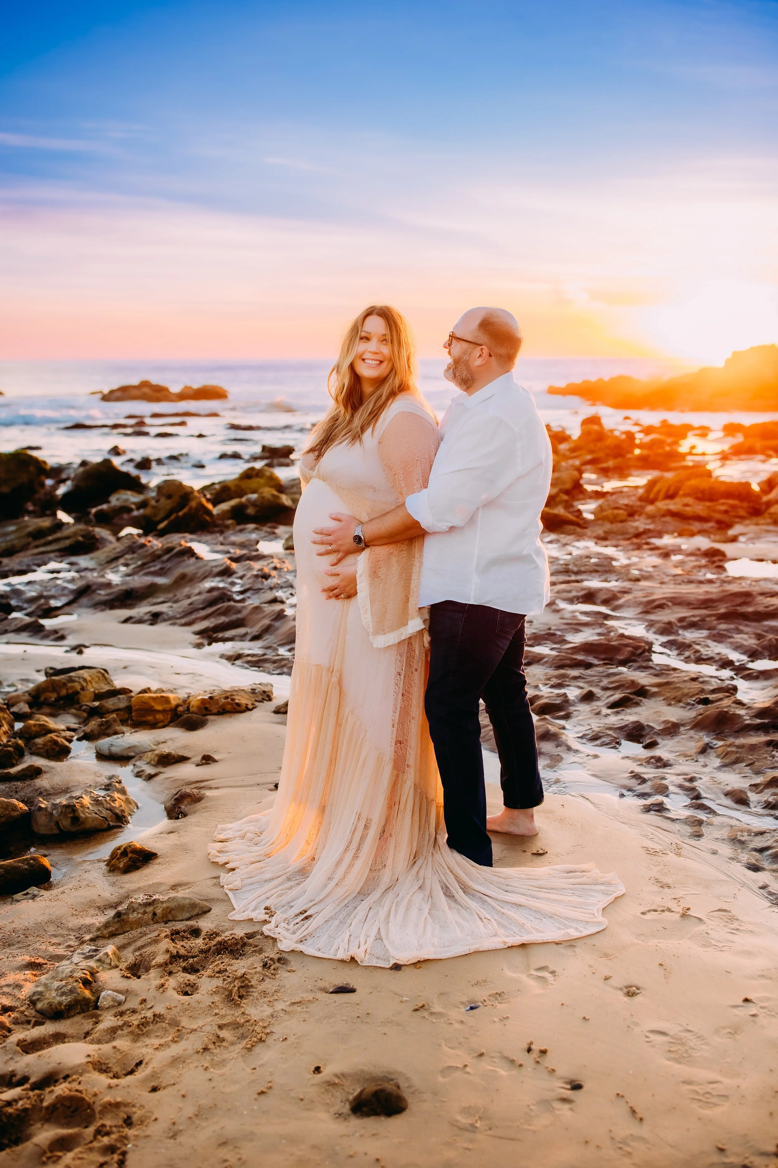 mom and dad to be smiling and holding her pregnant belly during their sunset beach photoshoot in Newport Beach, CA