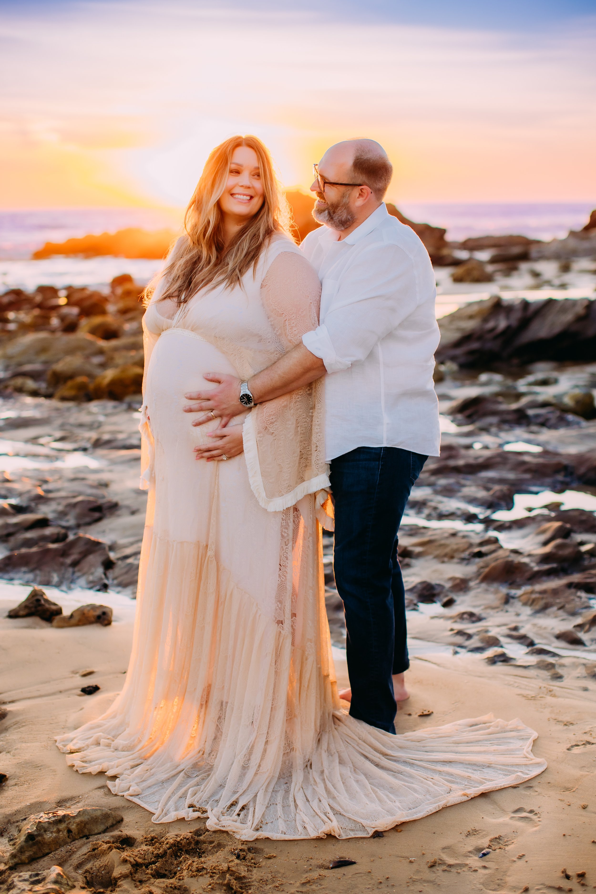 huband and wife laughing together with their hands on wife's pregnant belly during their sunset photo session in Newport Beach, Orange County, California