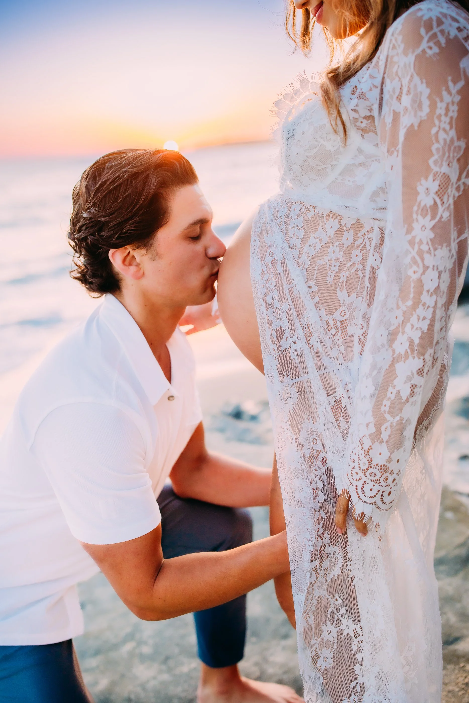 husband kissing his pregnant wife's belly as the sun sets behind them during their photoshoot in Laguna Beach, California