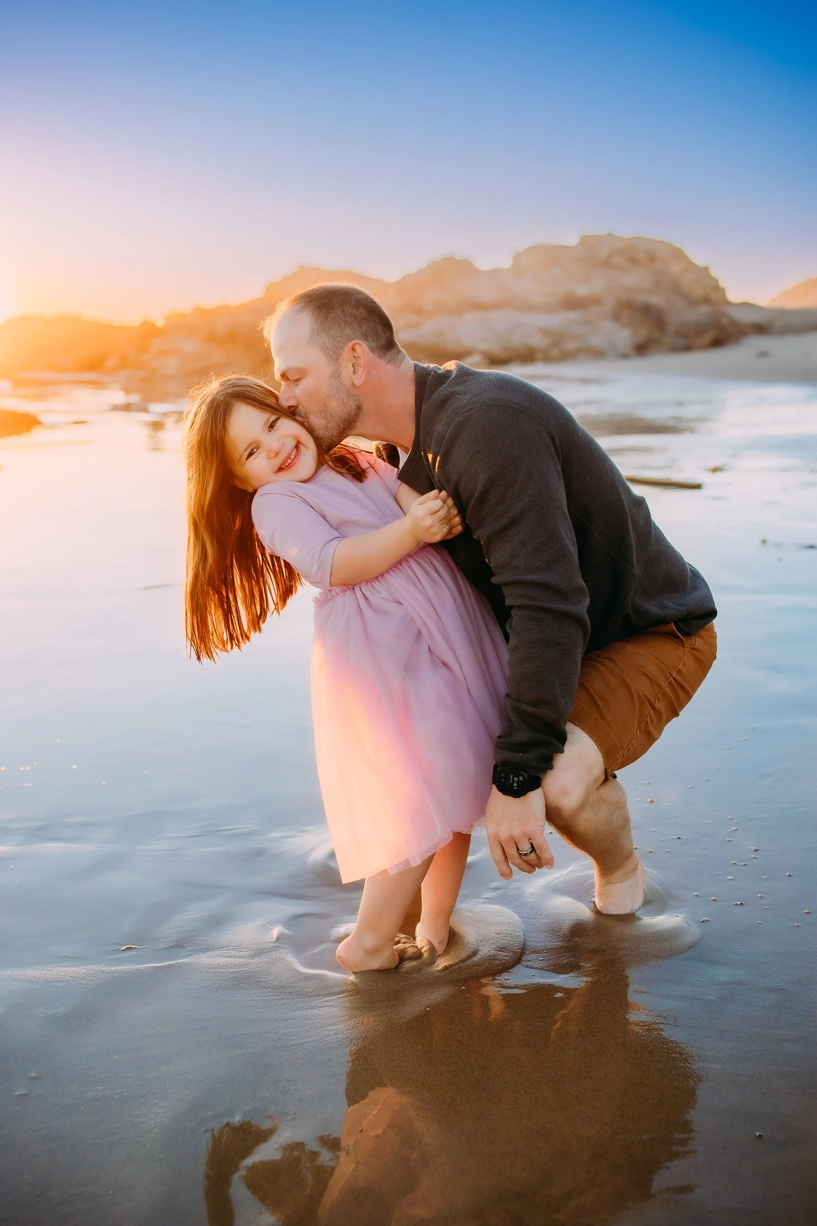 dad giving a kiss to his giggling daughter during while the sun sets during their family photo session in Newport Beach.