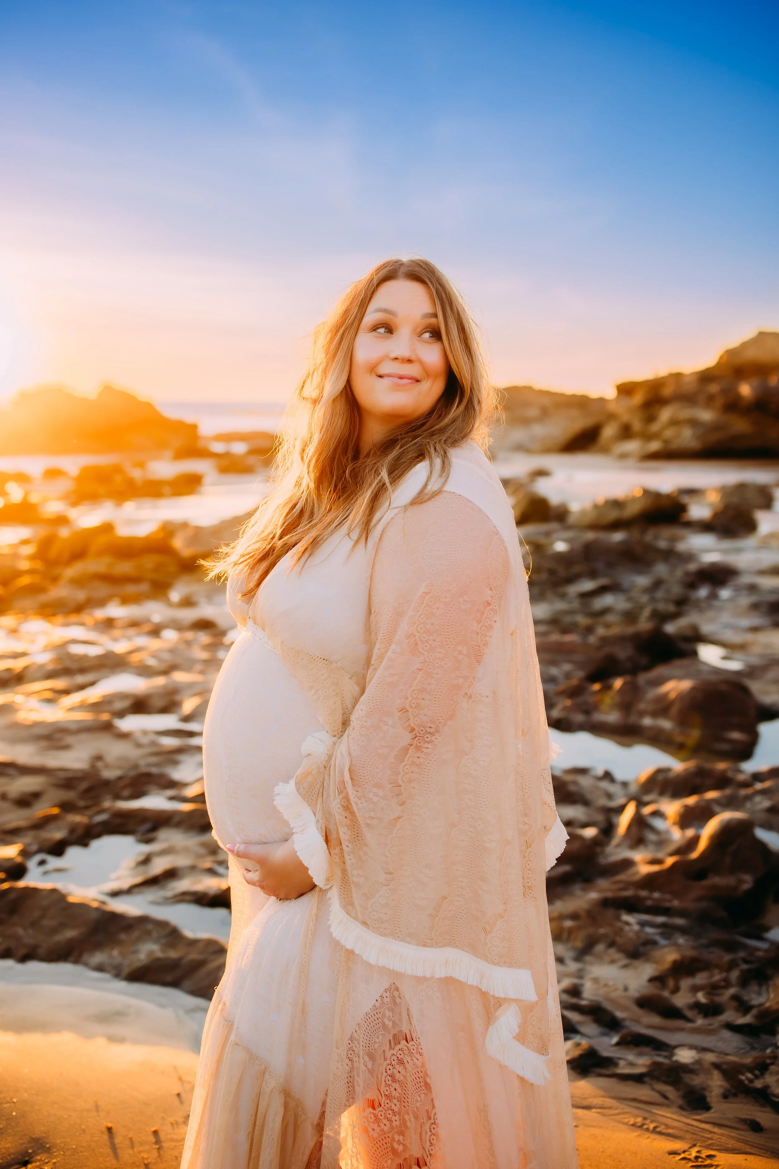 mom to be holding her belly as she gazes off with the sun setting behind her during her photo session in Newport Beach, CA