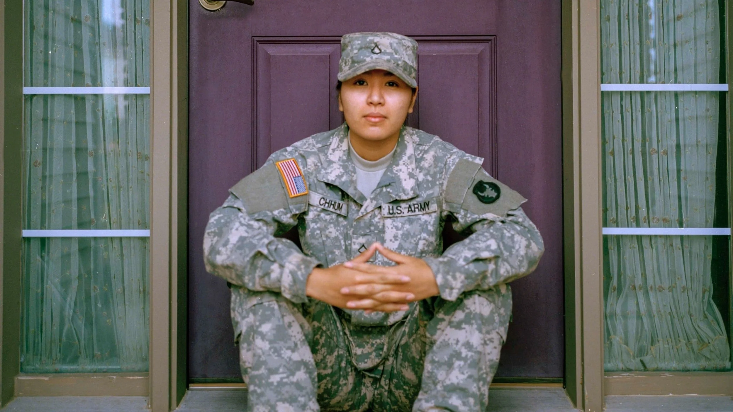Image of a member of the US army sitting on a doorstep in front of a purple door with hands clasped, wearing fatigues