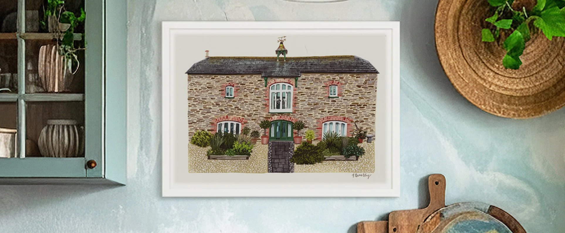 Framed illustration of a stone house with a dark roof, surrounded by plants and flowers, hanging on a light green wall.