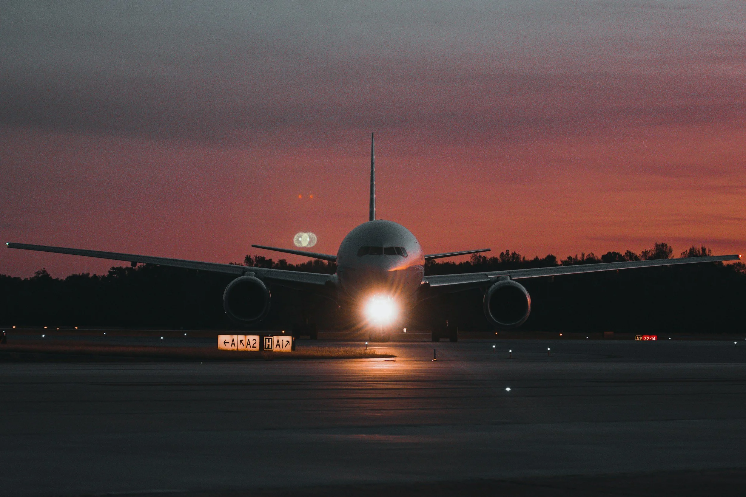 Commercial airplane on the runway at dusk, facing forward with lights on, dark silhouette of trees in the background, pinkish-orange sunset sky, runway lights visible.