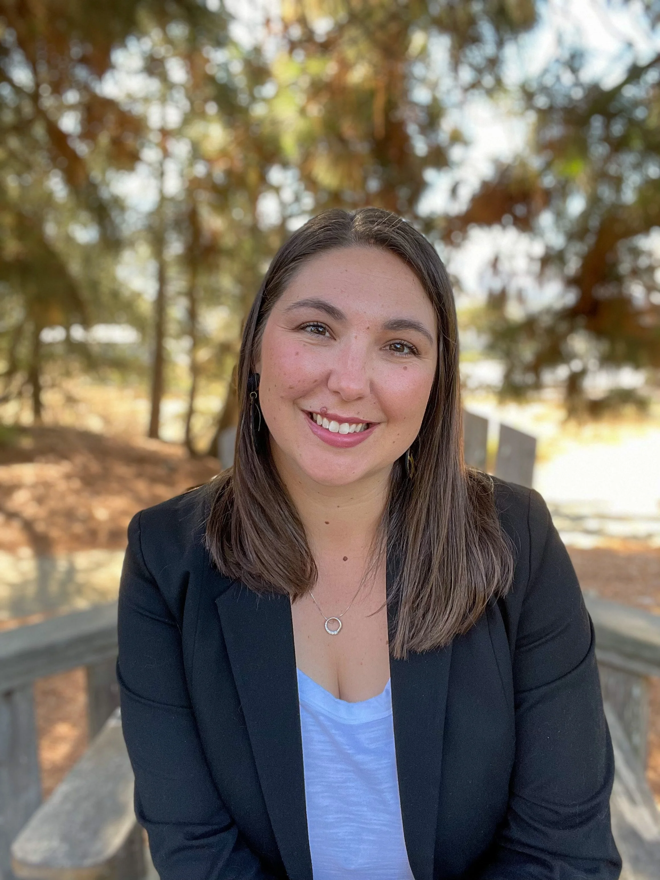 A woman with long brown hair smiling, wearing a black blazer and white top, sitting outdoors with trees and fall foliage in the background.