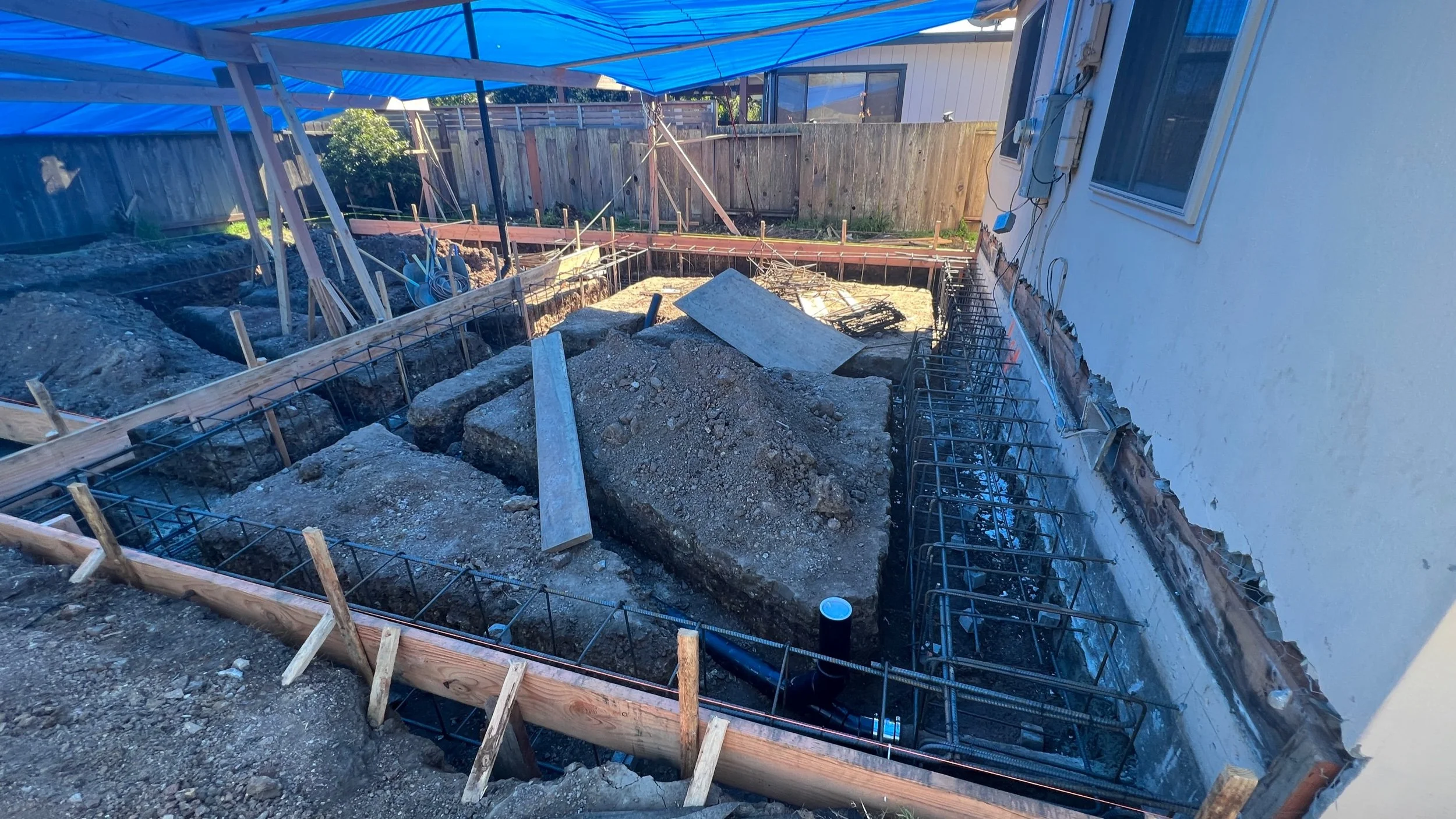 The ground is being excavated to match the required foundation dimensions. A rebar cage is then dropped in & concrete will be poured. Underground plumbing also needs to be planned prior to the concrete pour. Note how the rebar also ties into the exis
