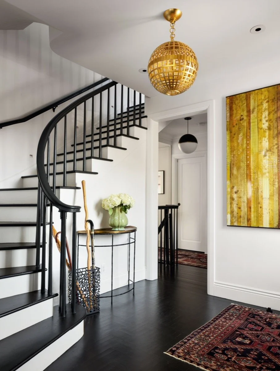  A white hall with a very dark wood floor and matching stair banister, with gold art, lighting and accents. 