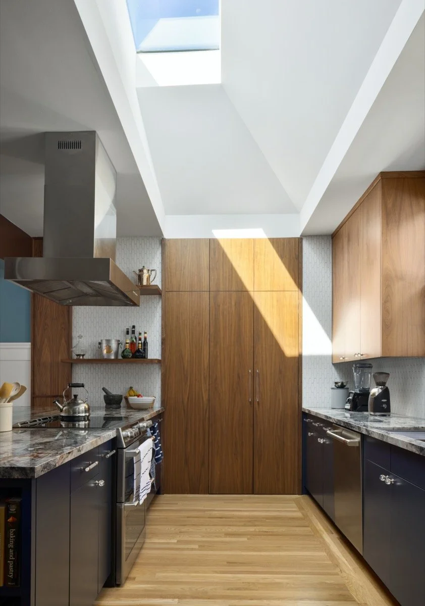  A cooktop on an island, with counters on the back wall, and a pantry on the right-side wall; a skylight lights the center of the kitchen. 