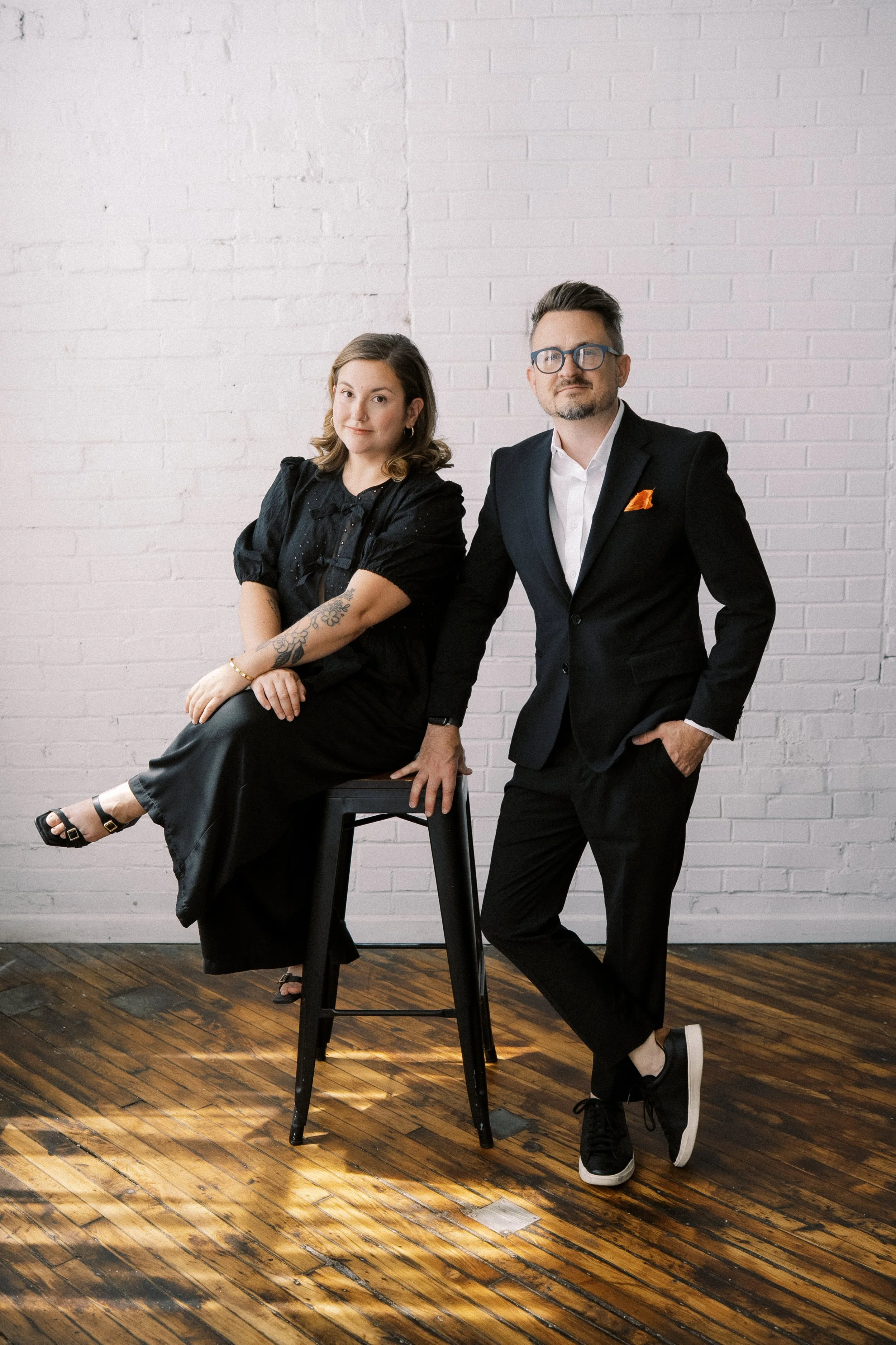 A man and woman pose in front of a white brick wall; the woman sits on a black stool with her legs crossed, wearing a black dress and sandals, while the man stands beside her in a black suit with a white shirt, casual black sneakers, and glasses.