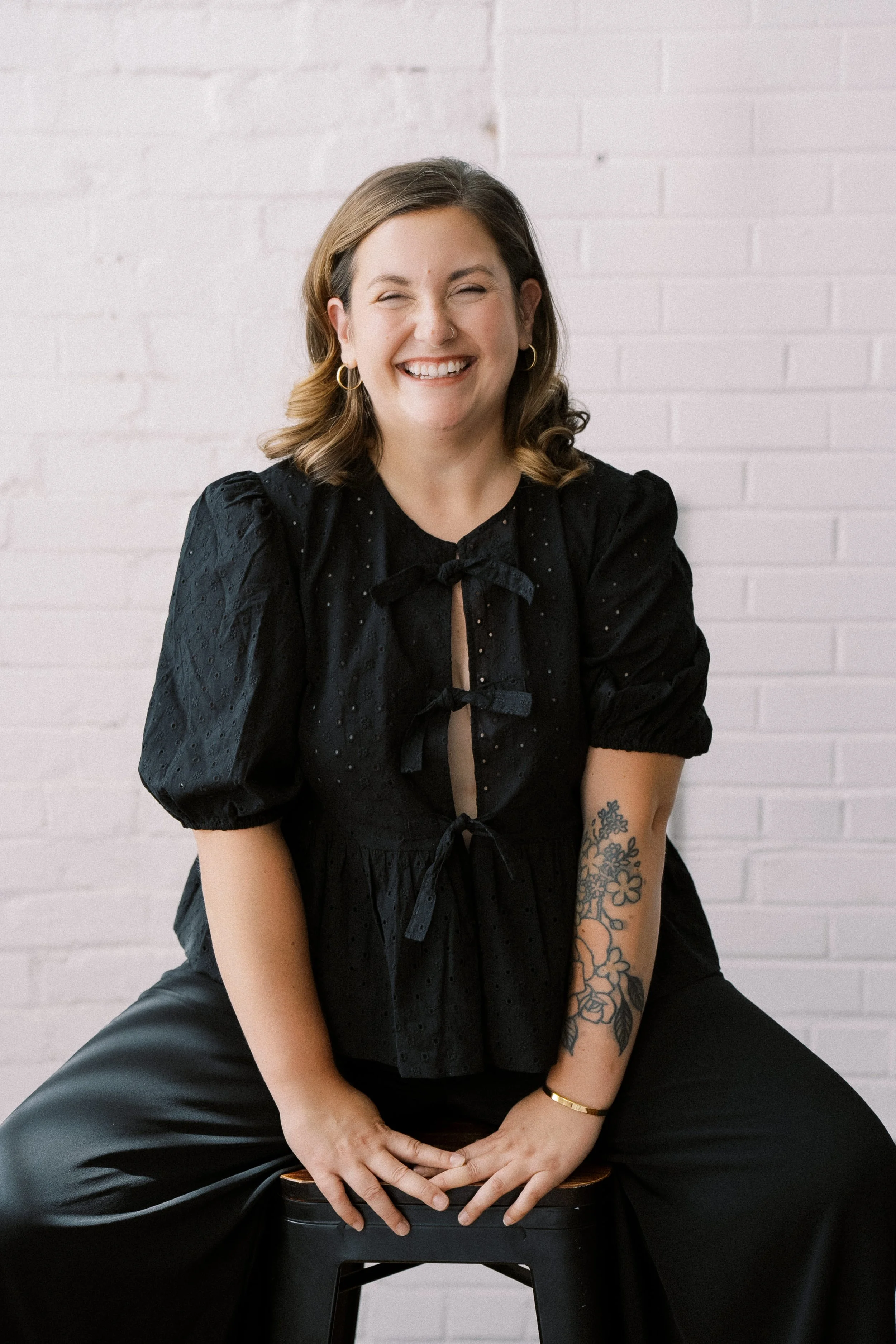 A woman with shoulder-length brunette hair, smiling and sitting on a black stool against a white brick wall. She is wearing a black puffy-sleeved blouse with a bow tie detail at the front, black wide-leg pants, hoop earrings, a bracelet, and has a fl