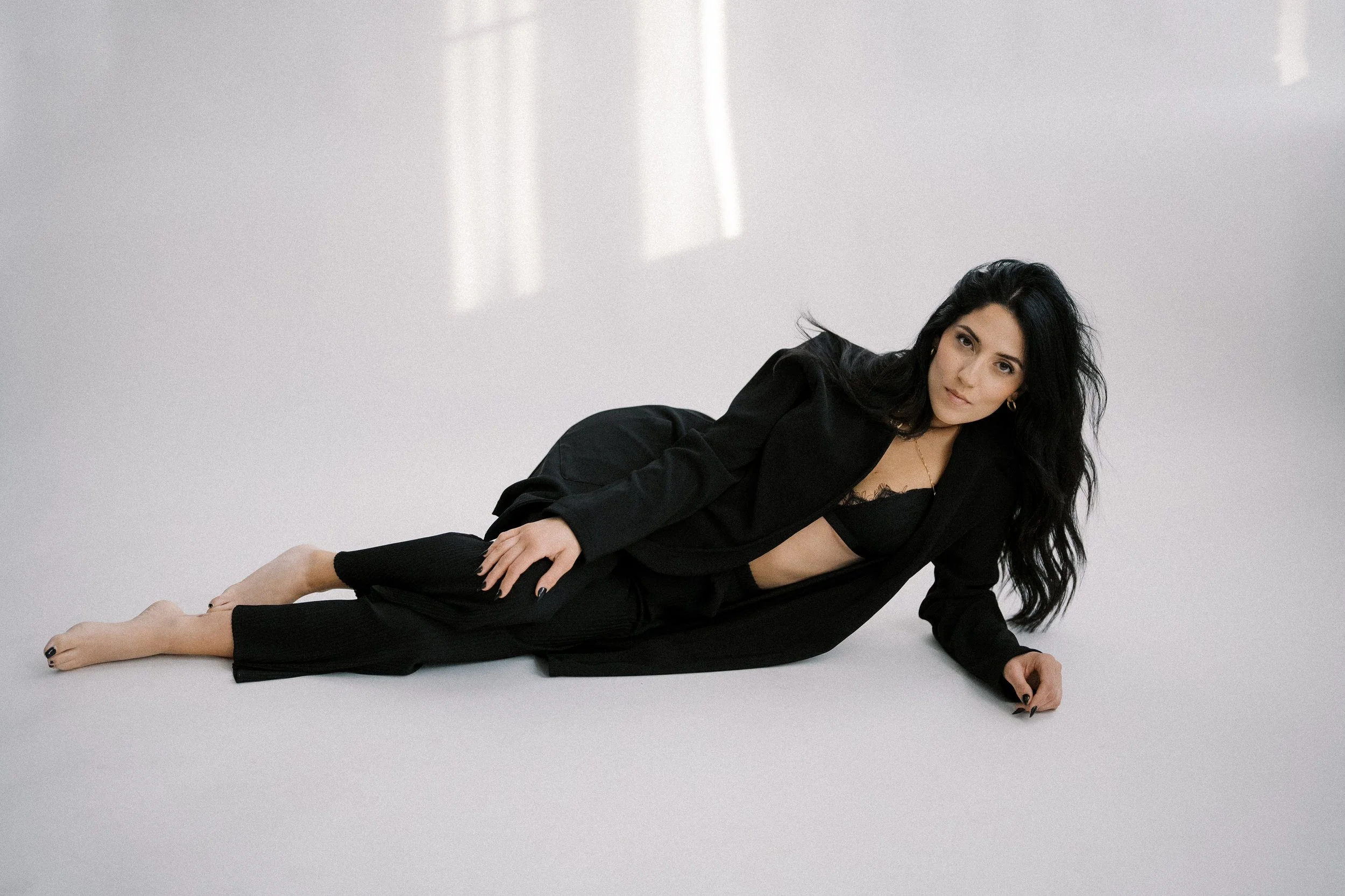 A woman with long black hair lying on her side on a white background, wearing a black blazer and black pants, with a black bra visible underneath.
