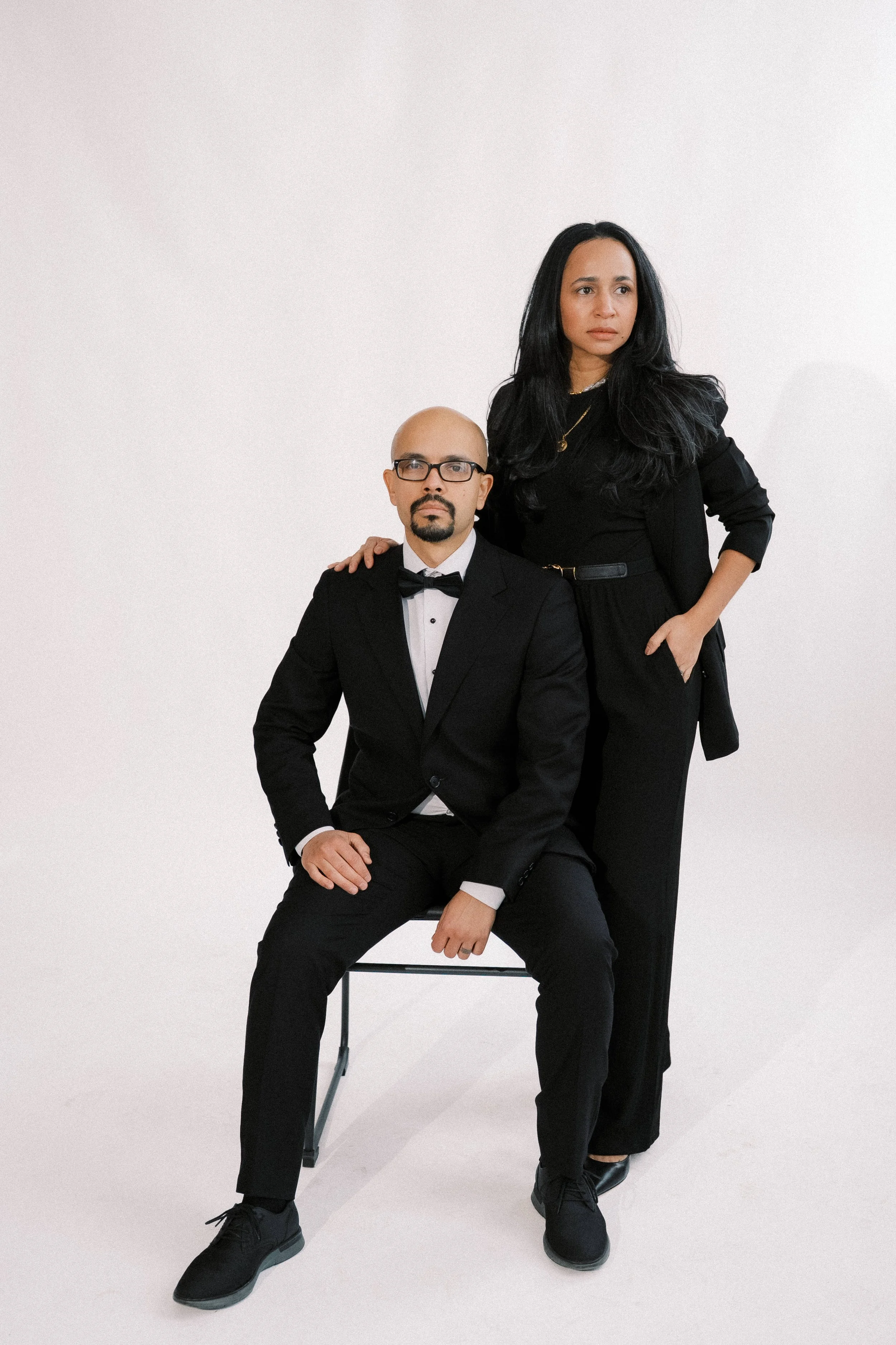 A man in a black tuxedo and glasses is sitting on a chair, with a woman in a black suit standing beside him, her hand on his shoulder, against a plain white background.