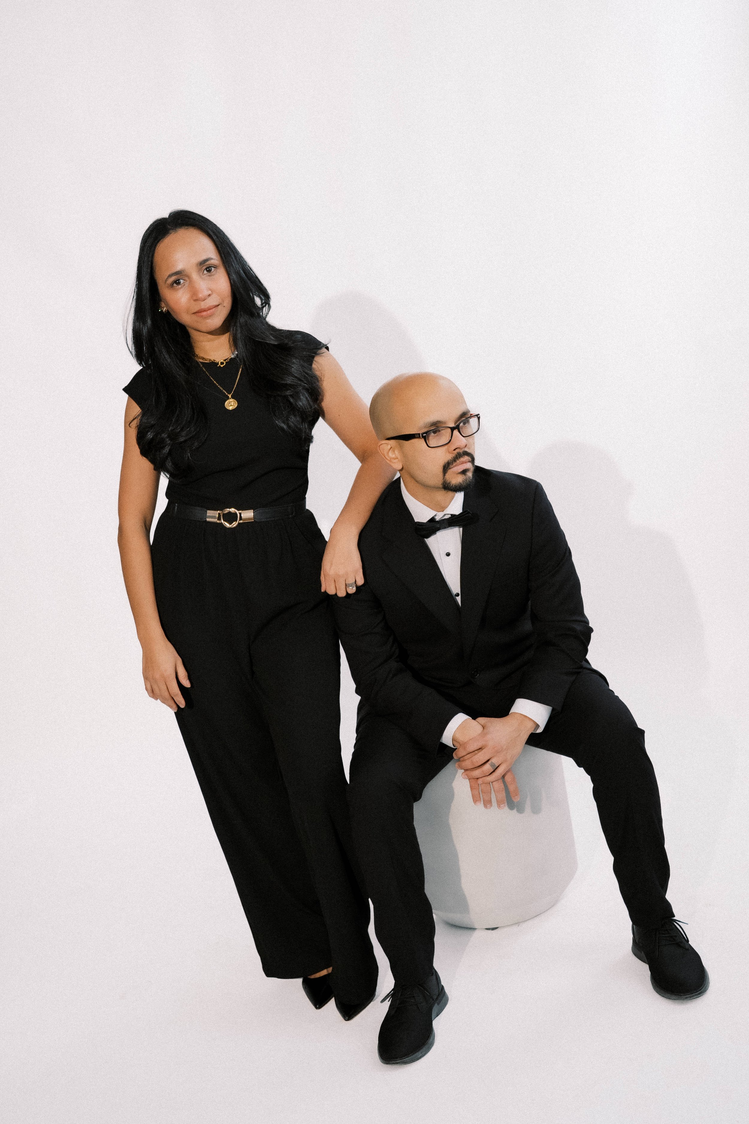 A woman in black standing with her hand on the shoulder of a man in formal wear, who is seated on a white stool against a plain white background.