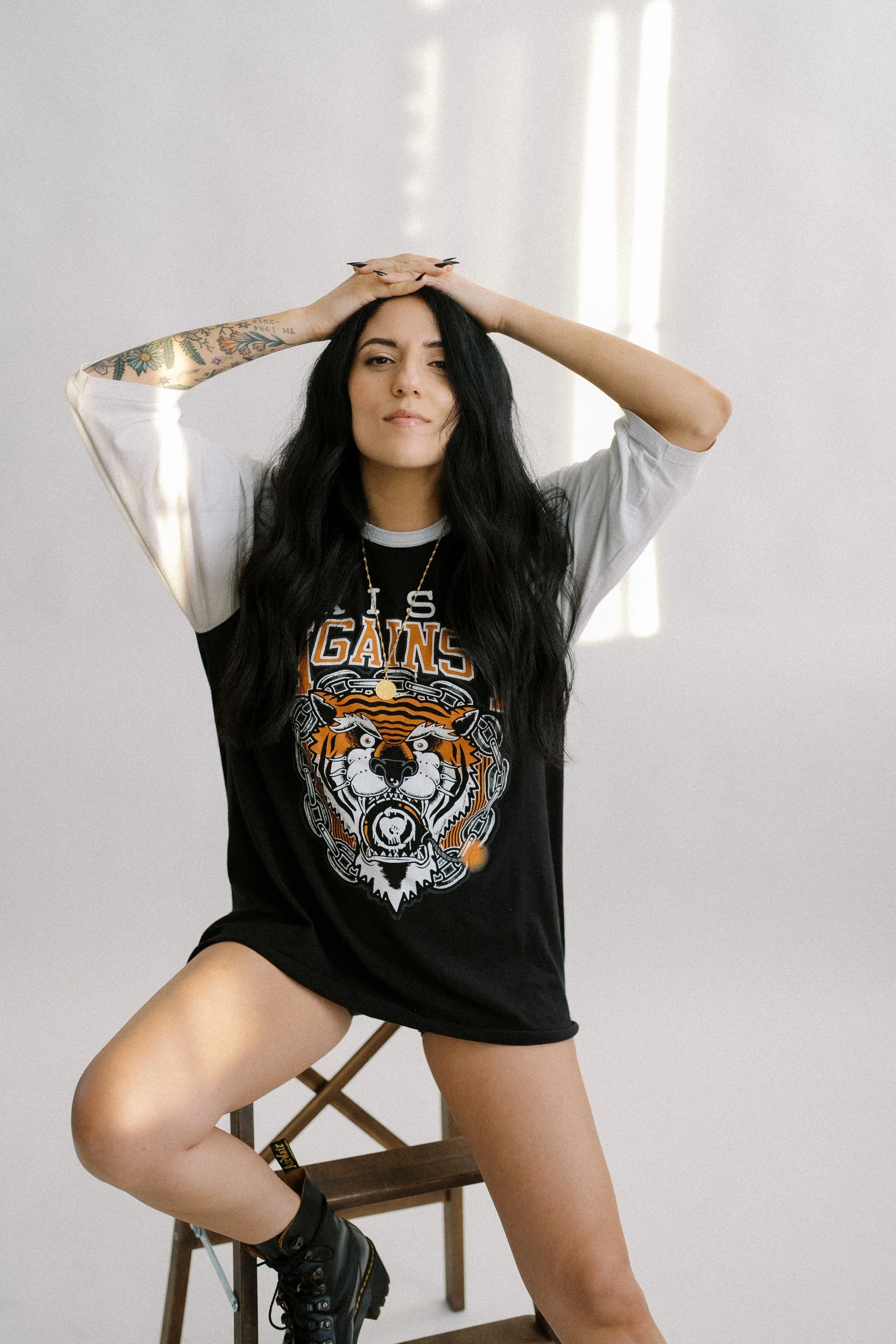 A woman with long black wavy hair and tattoos on her left arm, posing in front of a plain white background, wearing a black and white T-shirt with a graphic tiger and the words 'Los Angeles Gangs', black combat boots, and a necklace, sitting on a woo