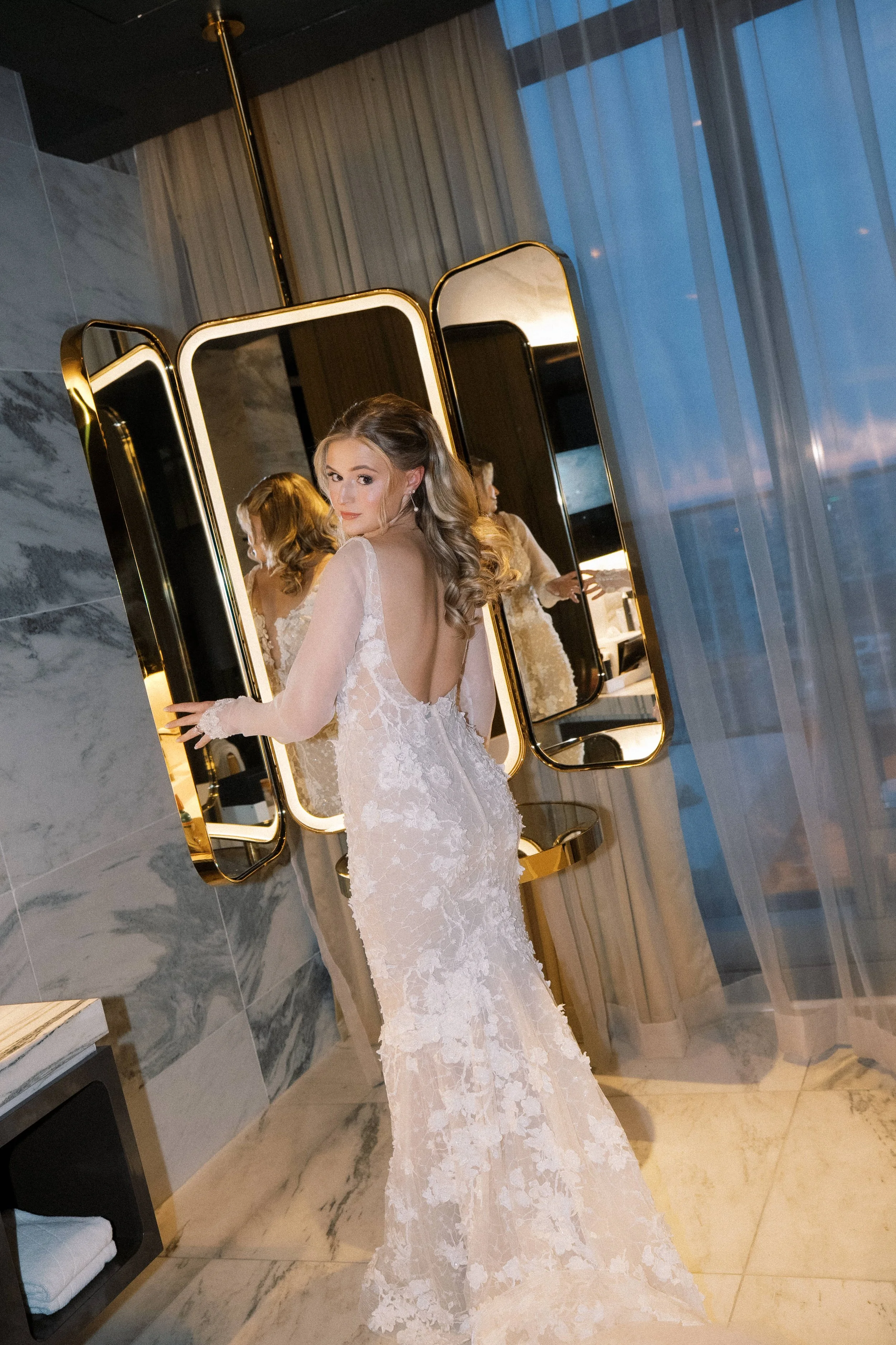 A woman in a lace wedding dress standing in front of three large mirrors in a modern hotel room, adjusting her dress and looking over her shoulder.