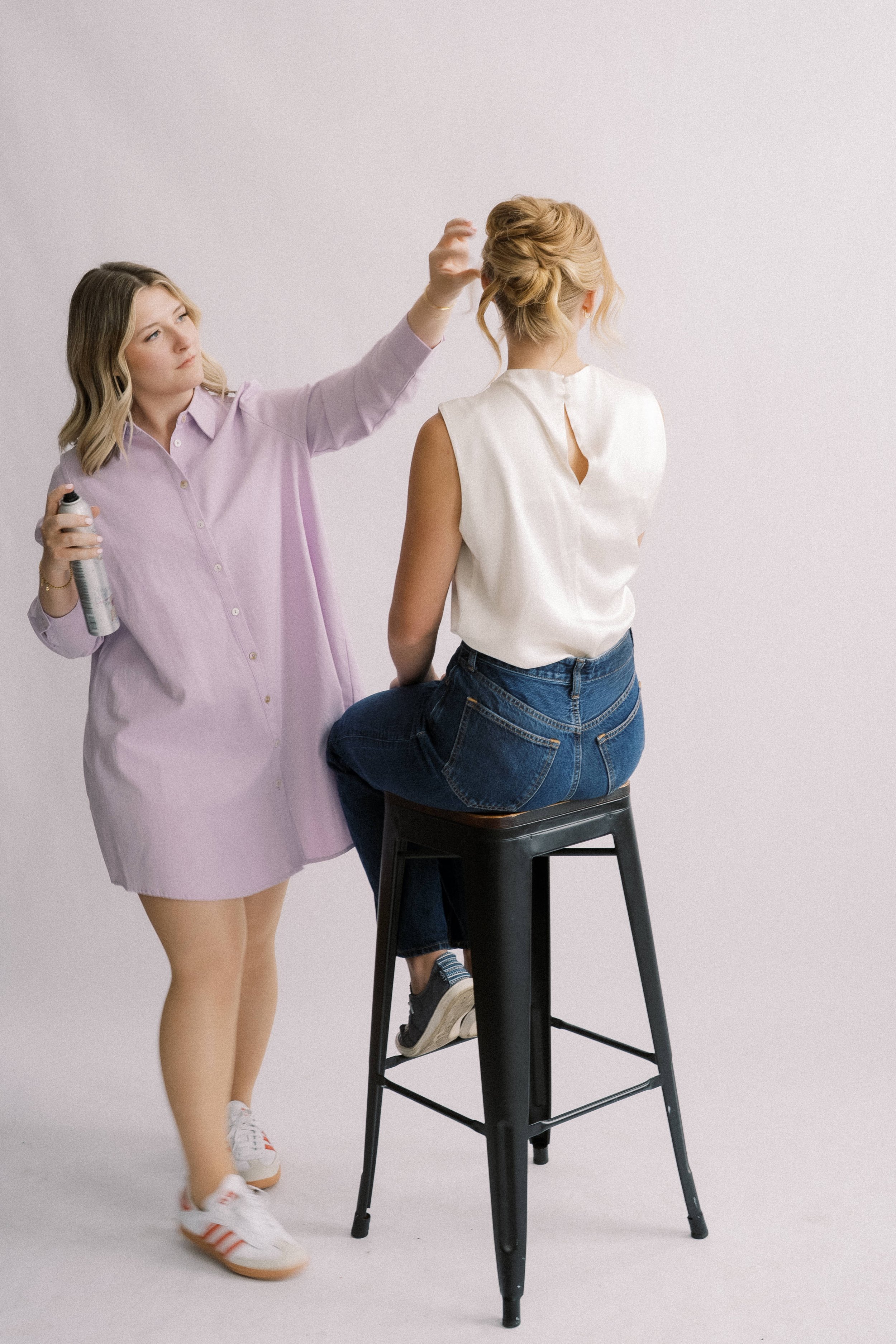 A woman sitting on a stool getting her hair styled by a stylist holding a hairspray can, against a plain white background.