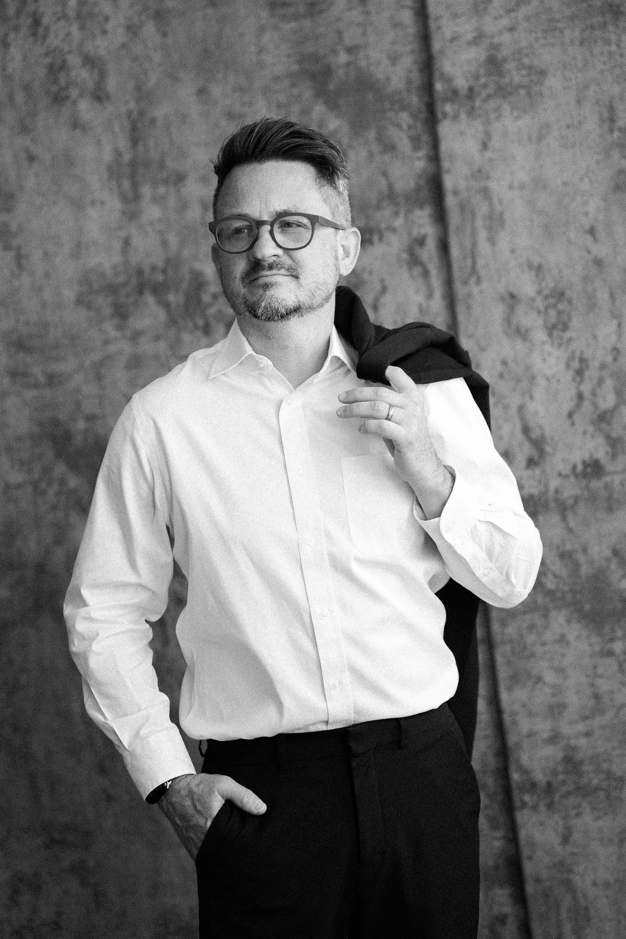 A man with glasses, a beard, and a mustache, wearing a white dress shirt and dark pants, standing against a textured wall. He has a jacket draped over his shoulder, held by his right hand, and his left hand is in his pocket.
