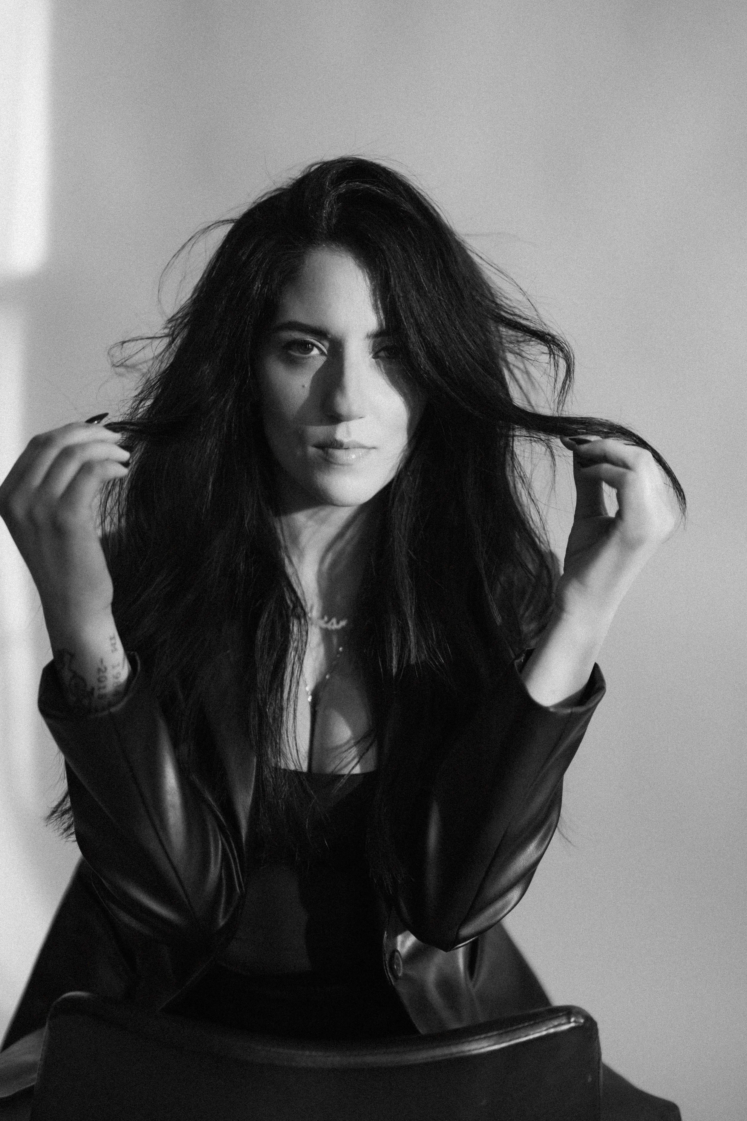 Black and white portrait of a woman with long, wavy dark hair, wearing a leather jacket, sitting against a plain background.