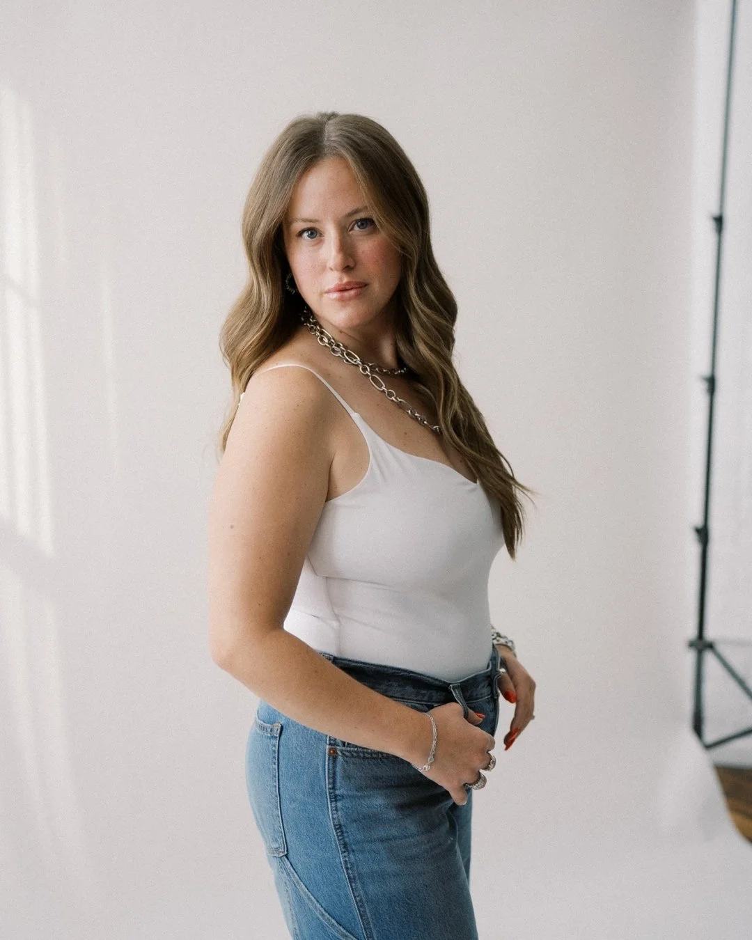 A woman with long, wavy brown hair wearing a white tank top, blue jeans, and silver jewelry standing indoors in front of a plain wall.