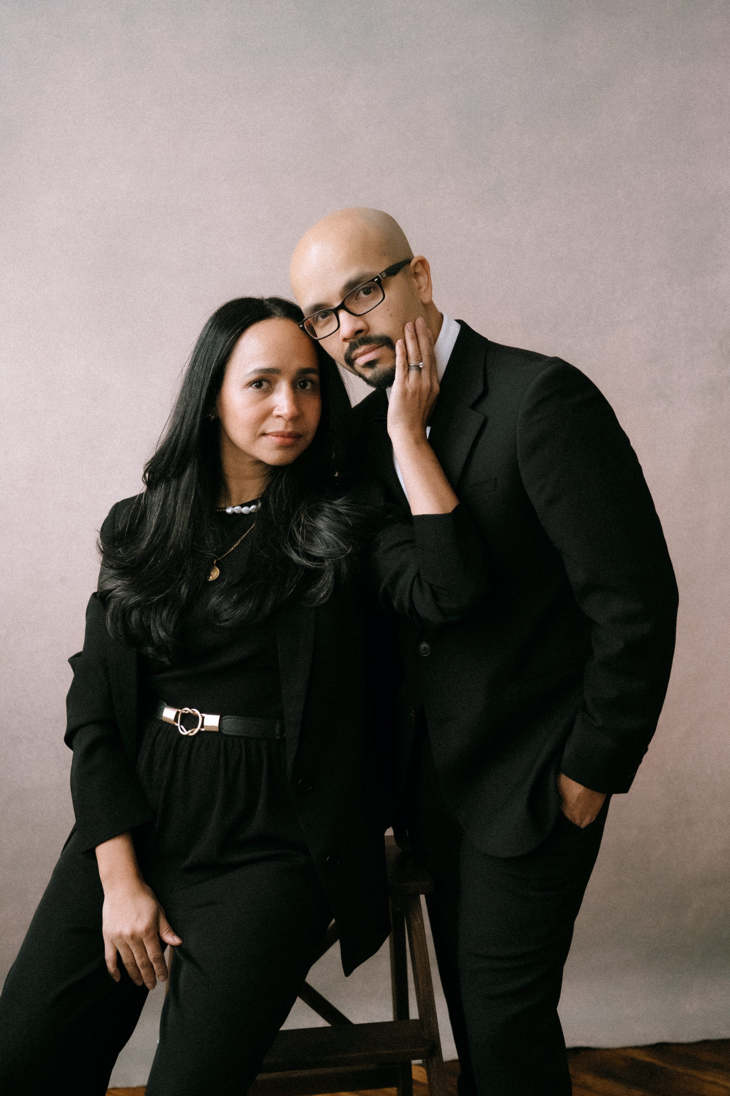 A man and woman dressed in black suits posing together indoors against a neutral background. The woman has long dark hair, and the man is bald with glasses. The woman is touching the man's face, and they are looking directly at the camera.