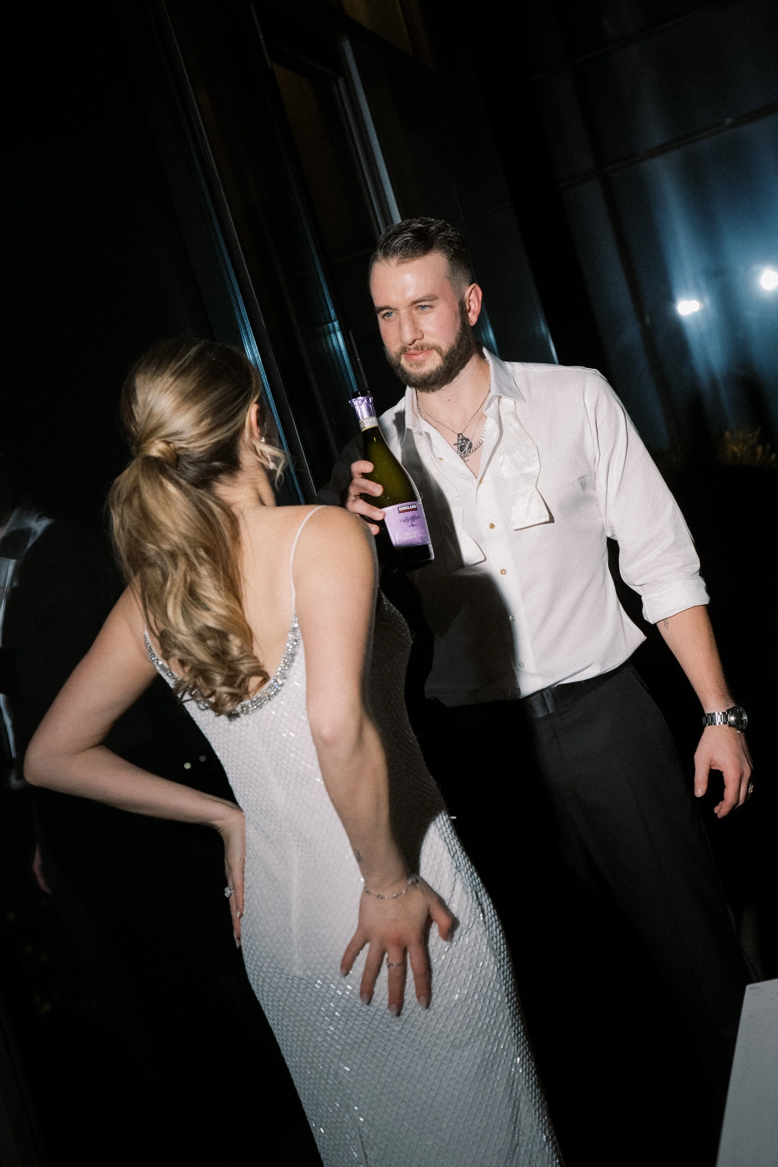 A young woman in a white sequin dress is talking to a man holding a bottle of drink at a nighttime social event.