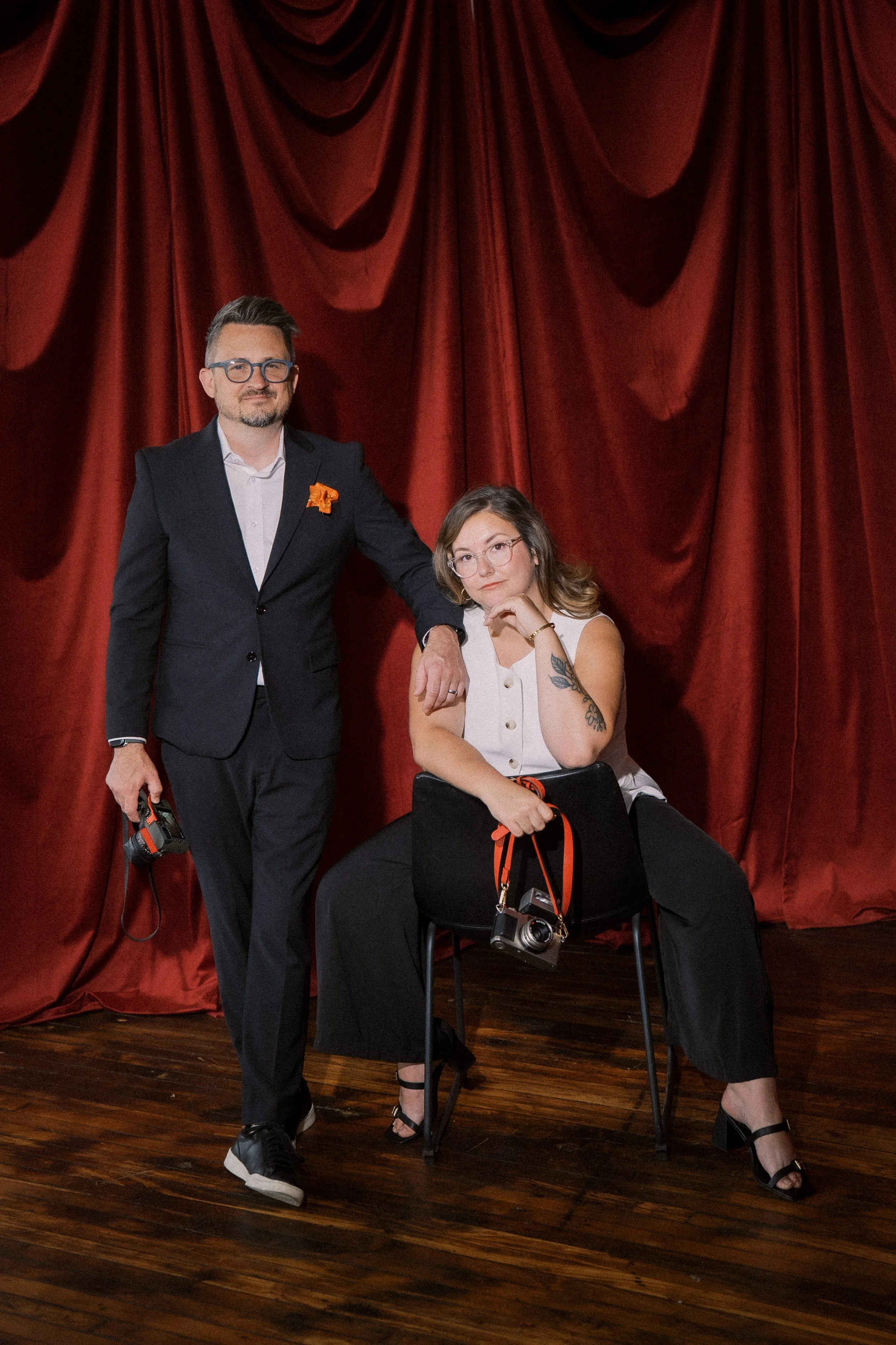 A man in a black suit standing next to a woman sitting on a chair, both against a red curtain backdrop, with cameras in hand, in a theater or stage setting.