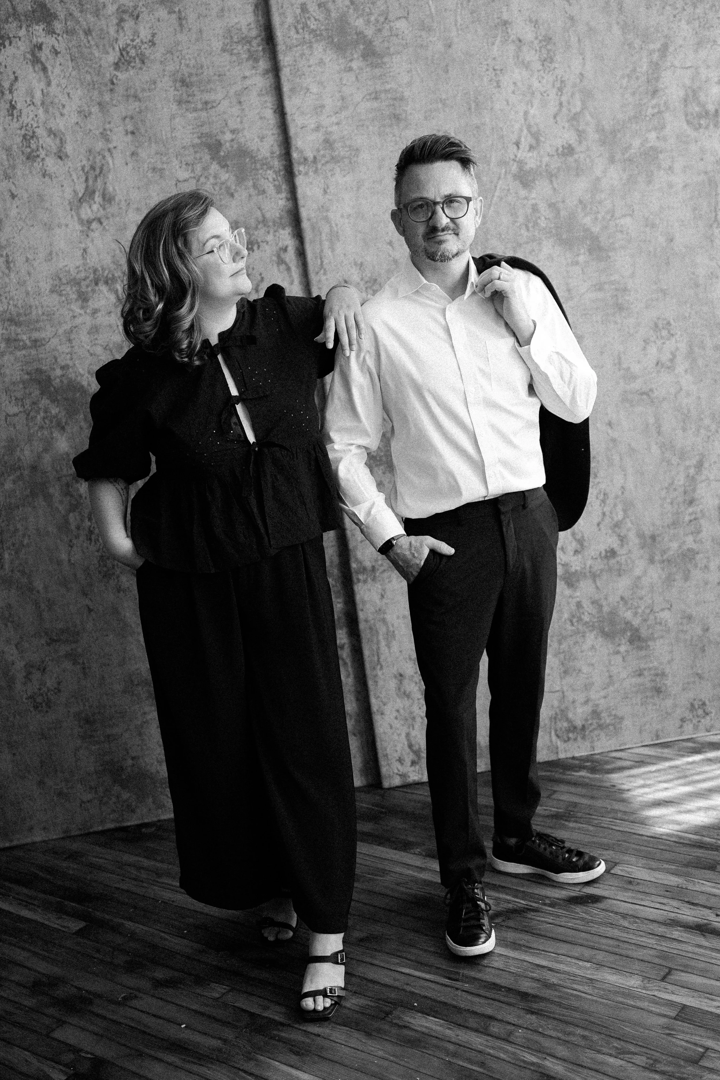 A black and white photo of a woman and a man standing together against a concrete wall. The woman is wearing a dark blouse and wide pants, with one hand behind her back and the other resting on the man's shoulder. The man is wearing glasses, a white 