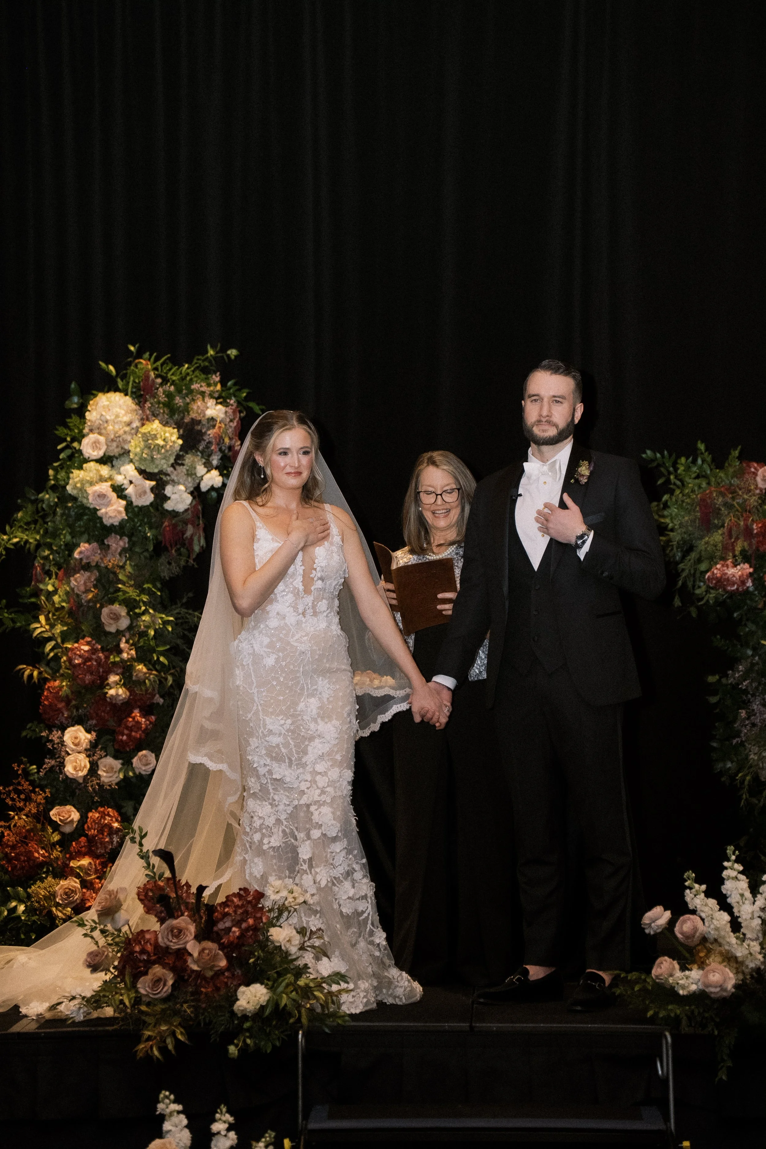 A bride and groom share a moment during their wedding ceremony, standing hand in hand in front of floral arrangements, with an officiant behind them, on a stage with a black curtain backdrop.