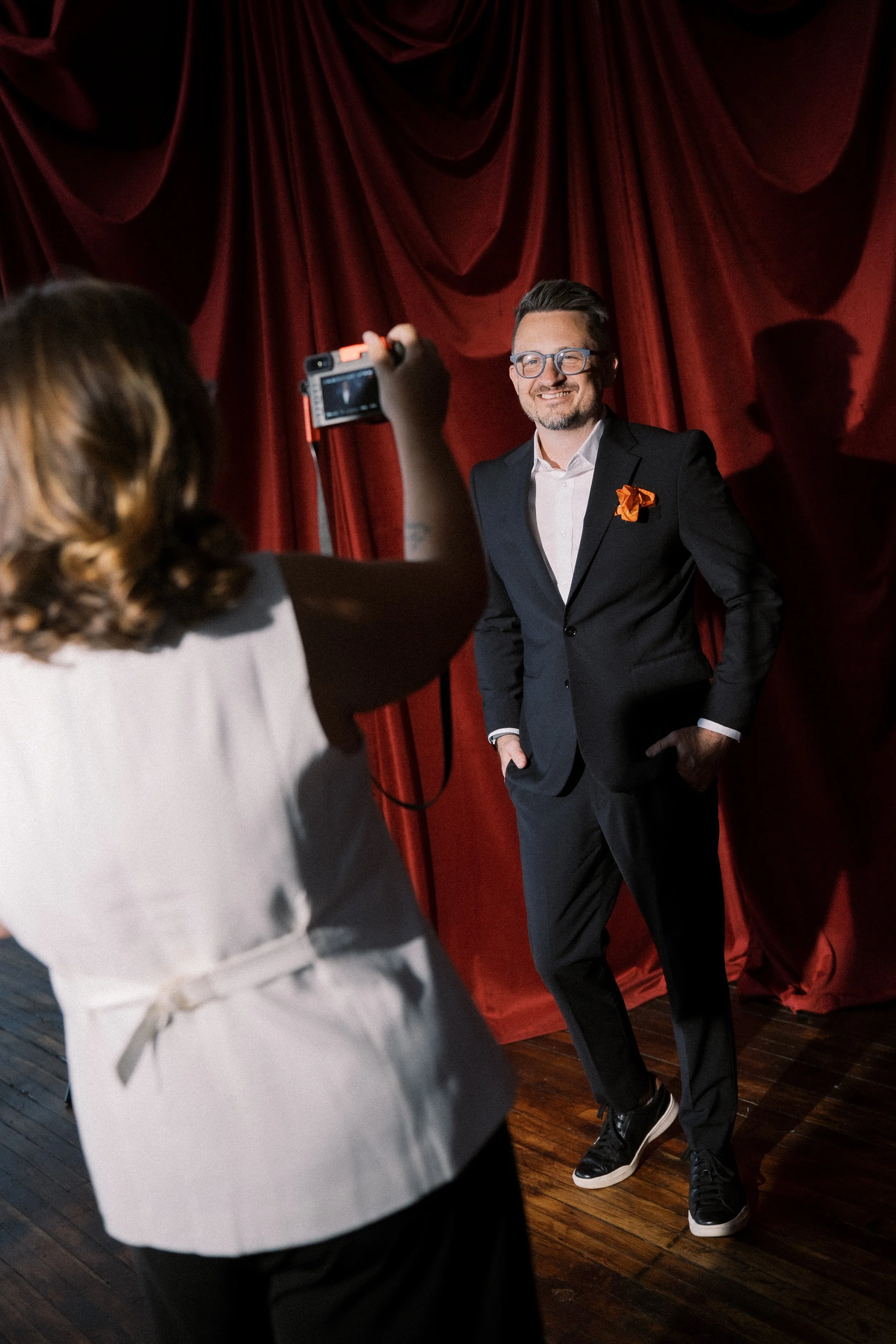 A woman is taking a photo of a man posing in front of a red velvet curtain. The man is wearing a black suit, white shirt, black sneakers, and glasses, smiling and standing with hands in pockets.