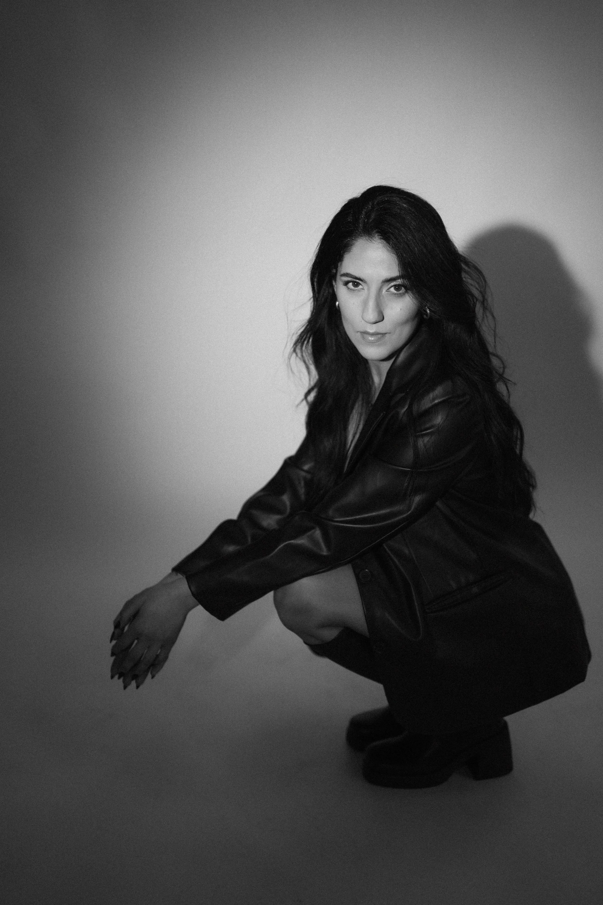 A black-and-white photo of a woman with long dark hair squatting and looking at the camera. She is wearing a leather jacket and platform boots.