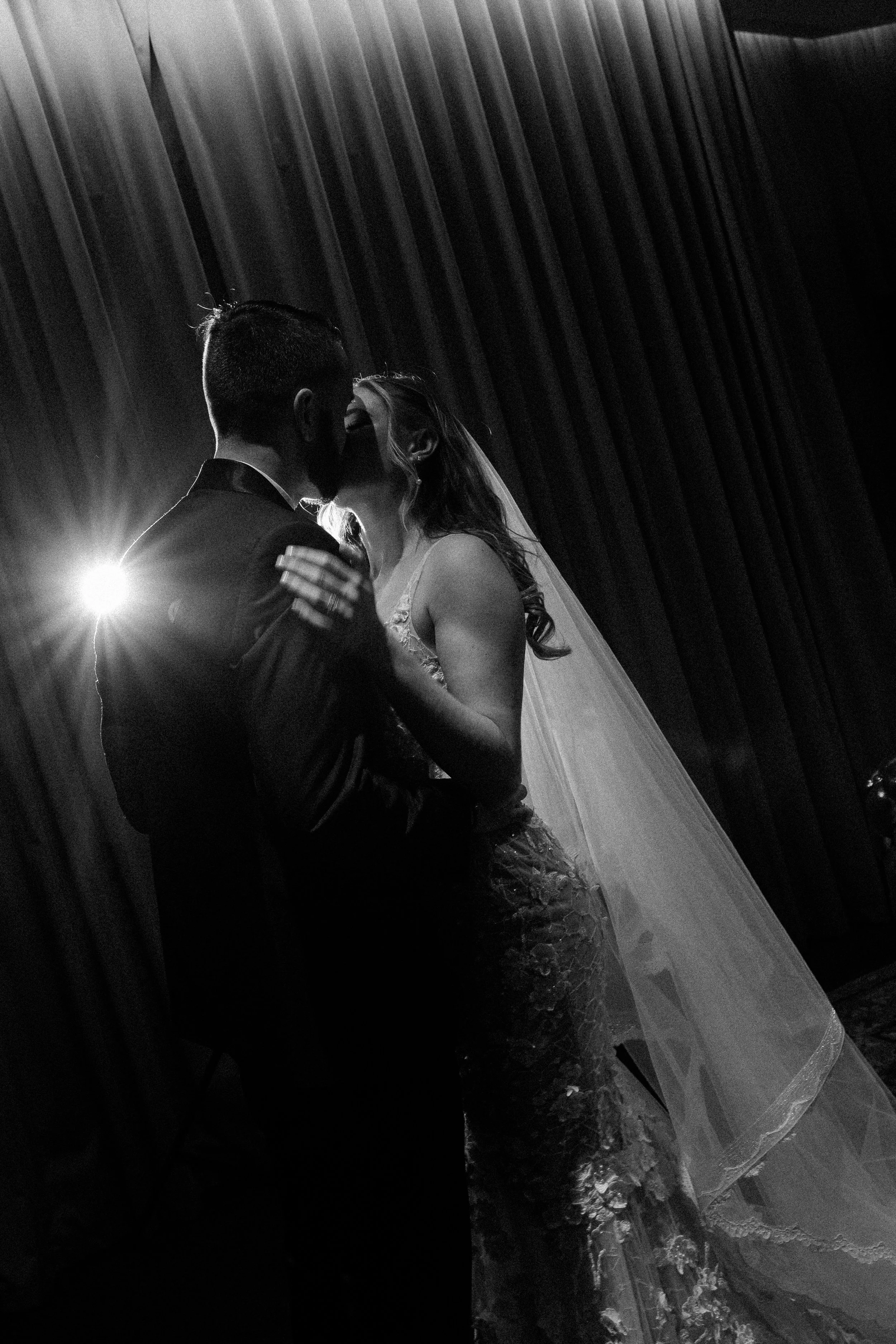 A couple in wedding attire sharing a kiss during their wedding reception with dramatic backlighting and curtain backdrop.