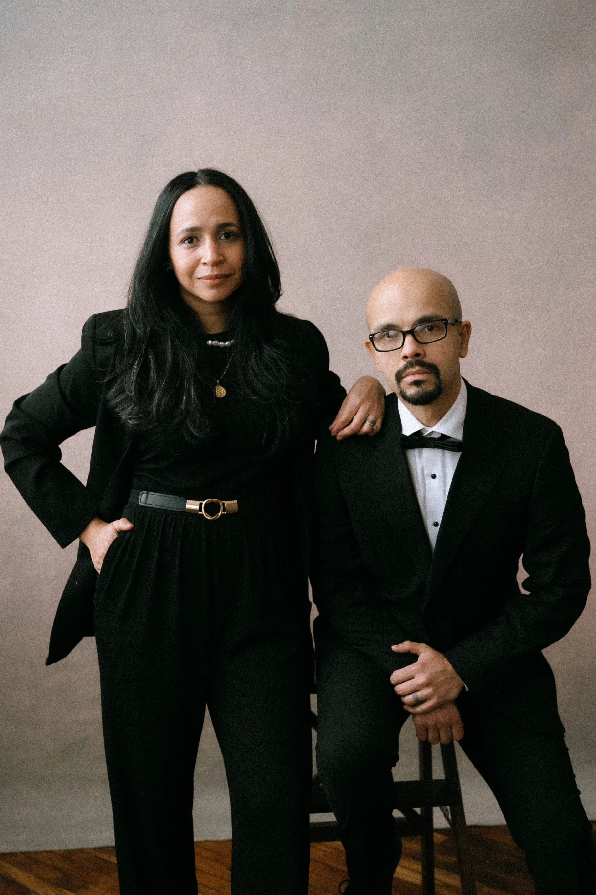 A woman with long dark hair in black clothing standing next to a man with a shaved head and glasses, dressed in a tuxedo, against a neutral background.