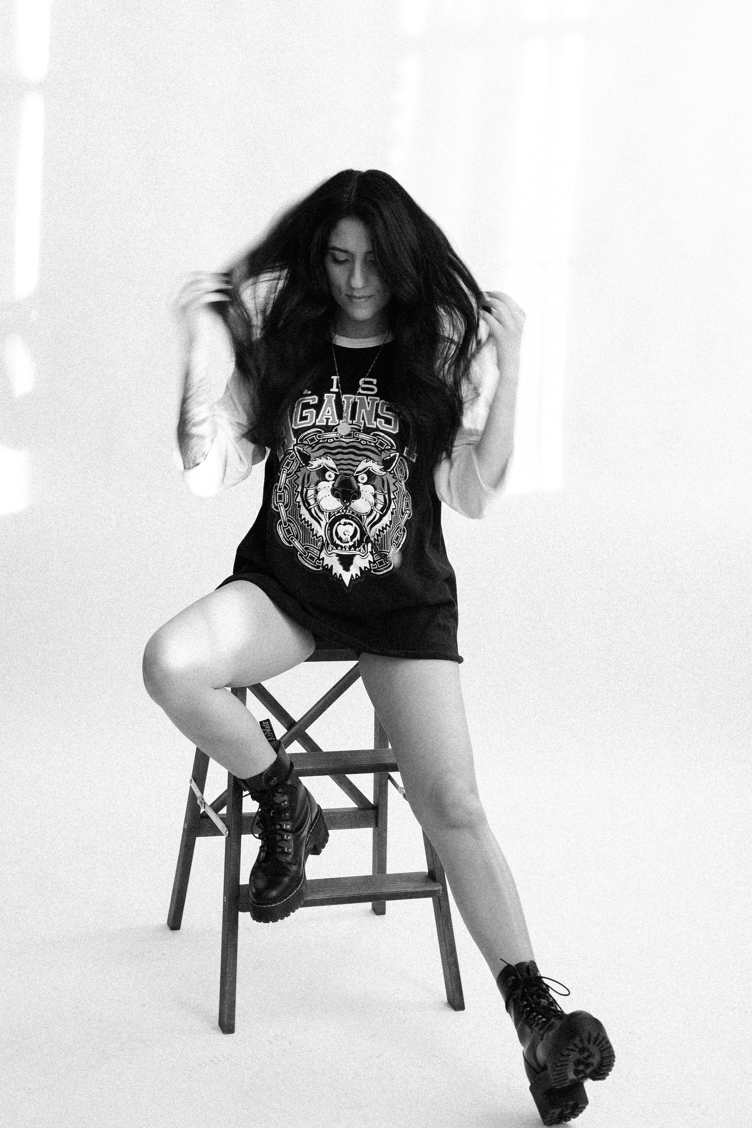 A woman with dark hair, wearing a graphic t-shirt, shorts, combat boots, and holding her hair, sitting on a wooden stool in a plain white studio background.