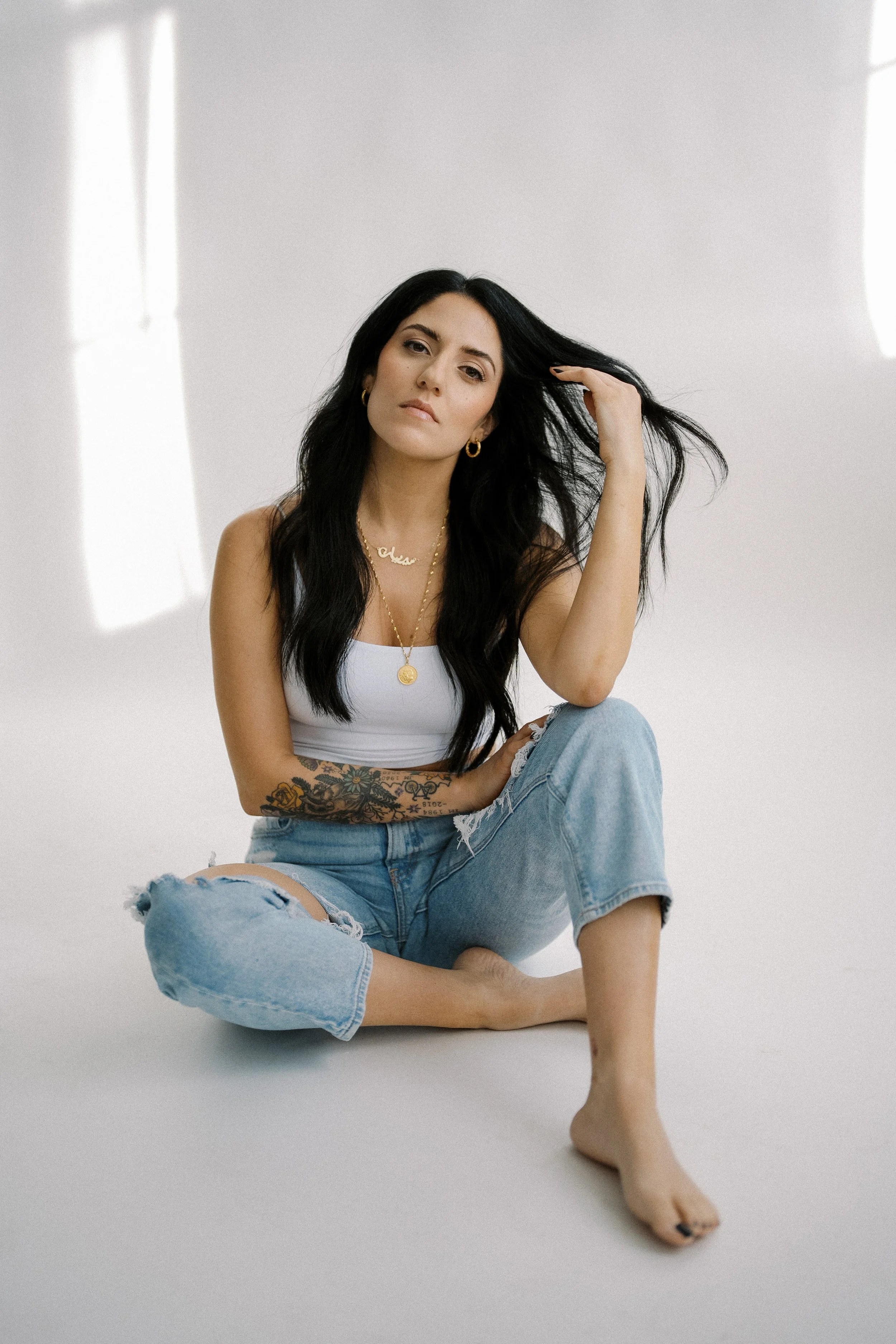 A young woman with long black hair, wearing a white tank top, ripped jeans, and gold jewelry, sitting barefoot on a plain white background. She has a tattoo on her left arm and is touching her hair.